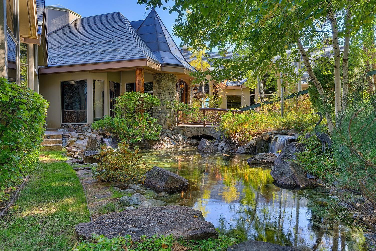 This image showcases a beautifully landscaped yard with a serene pond as its centerpiece. The pond features clear water, large rocks, and small waterfalls, reflecting the surrounding trees and the architecture of the house. A charming bridge leads to the house, which has a unique turret-style roof and stone accents, creating a fairytale-like ambiance.