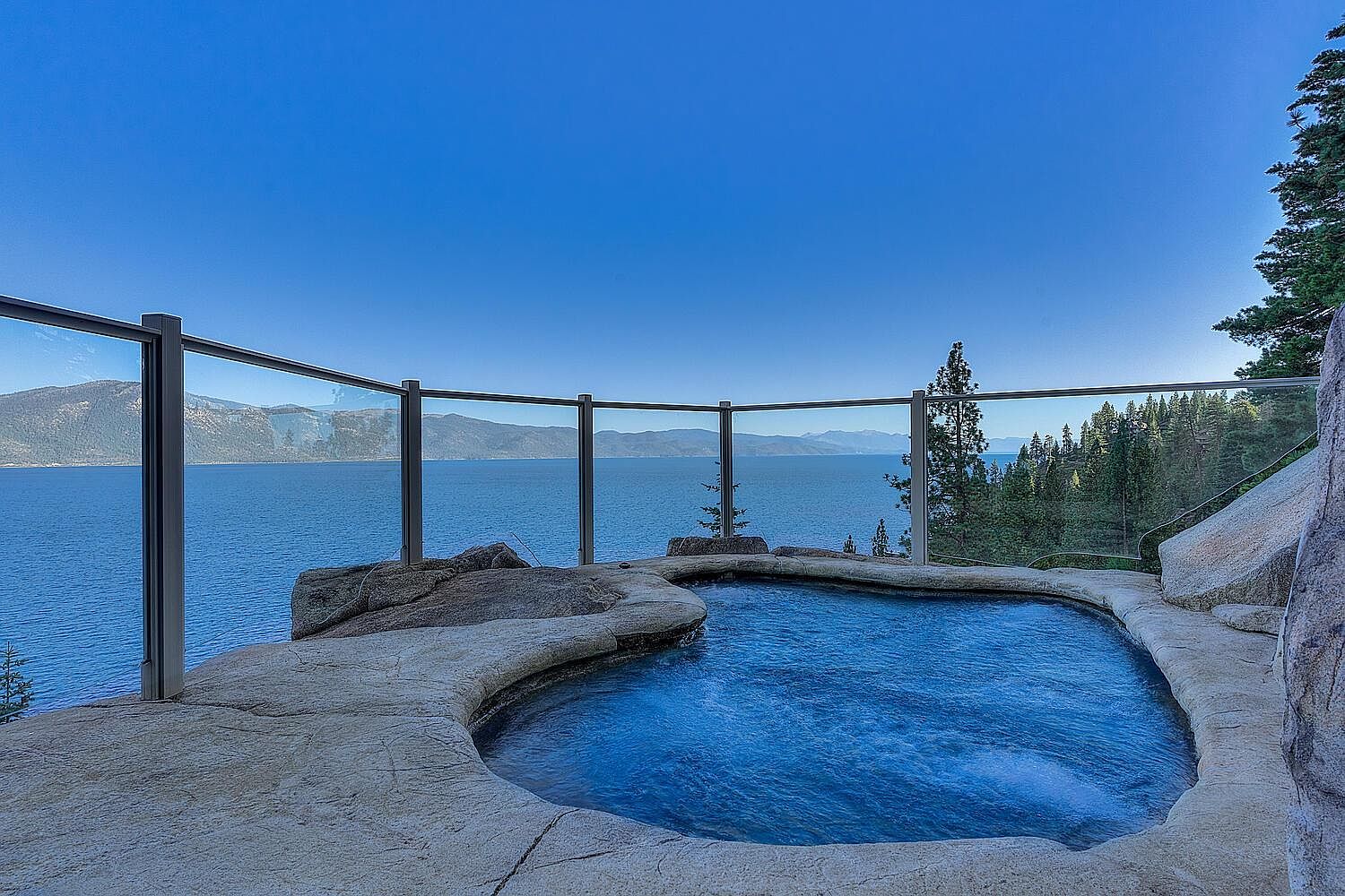 This image showcases an outdoor spa with a stunning view of a large body of water and distant mountains. The spa is constructed with natural stone, blending seamlessly into the landscape. A glass barrier provides safety without obstructing the panoramic vista, creating a luxurious and serene atmosphere.