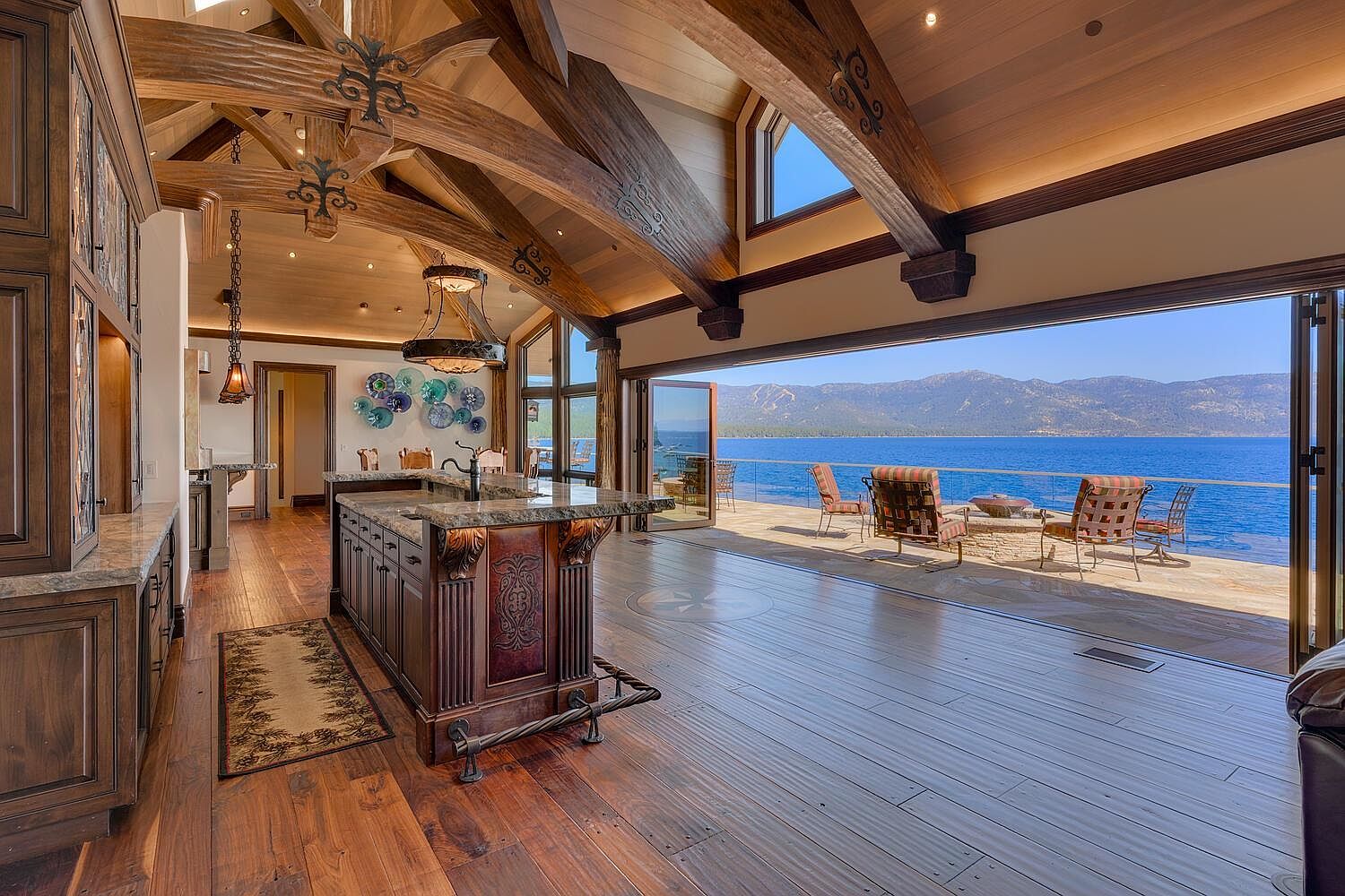 This interior shot showcases a luxurious kitchen with rich wood cabinetry, granite countertops, and an expansive island. The open floor plan seamlessly connects the kitchen to an outdoor patio with stunning lake views, creating an inviting indoor-outdoor living space. Exposed wooden beams and ornate lighting fixtures add to the room's character and upscale appeal.
