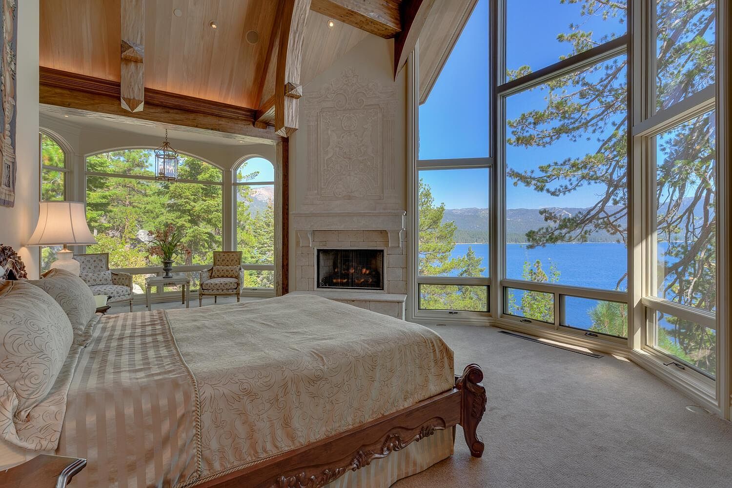 This is a luxurious primary bedroom featuring a large bed with patterned bedding, a stone fireplace, and expansive windows offering stunning views of a lake and surrounding trees. The room has a high, wood-paneled ceiling and elegant architectural details, creating a serene and upscale atmosphere. The room also features two chairs and a small table near the windows.