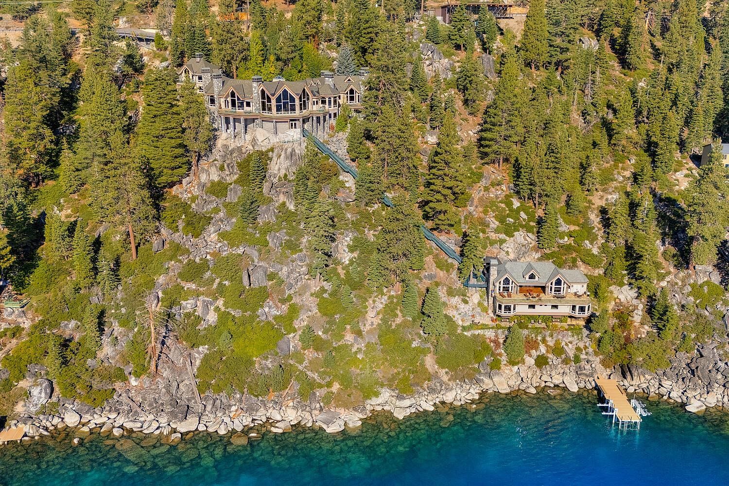 This aerial view showcases two luxury homes perched on a rocky cliffside overlooking a pristine blue lake. The larger home features multiple gables, stone accents, and a funicular for access, while the smaller home boasts a spacious deck and private dock. Lush greenery surrounds both properties, creating a sense of seclusion and natural beauty.