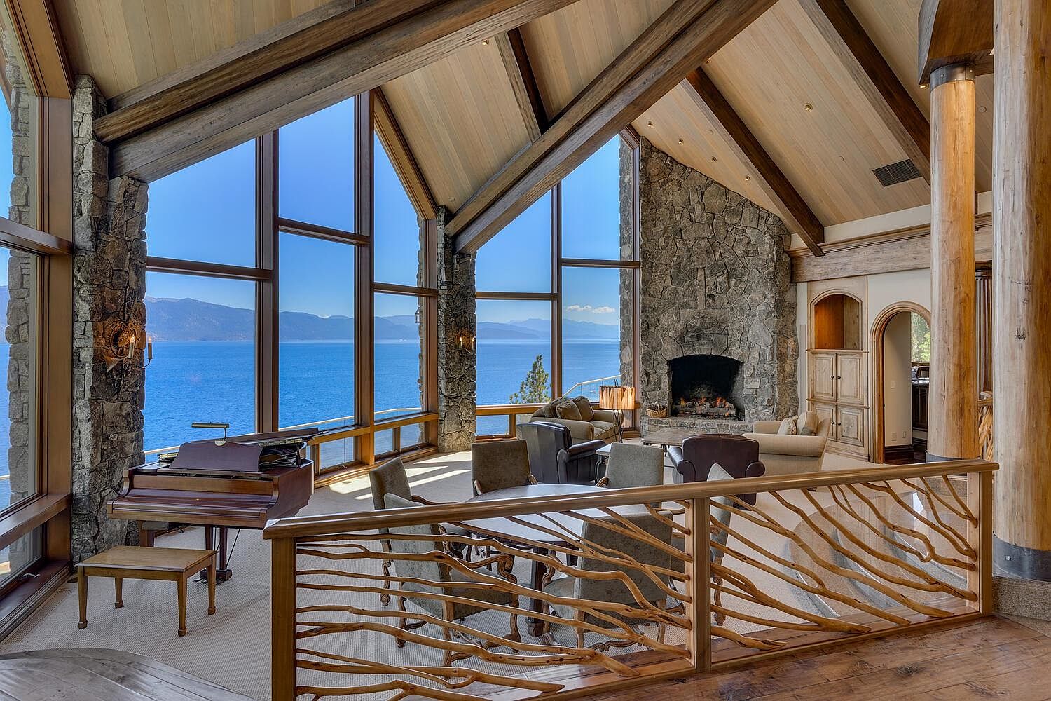 This is a luxurious living room with a stunning view of the lake through large windows. The room features a stone fireplace, wooden beams, and a grand piano, creating a warm and inviting atmosphere. The unique wooden railing adds a rustic touch to the elegant space.