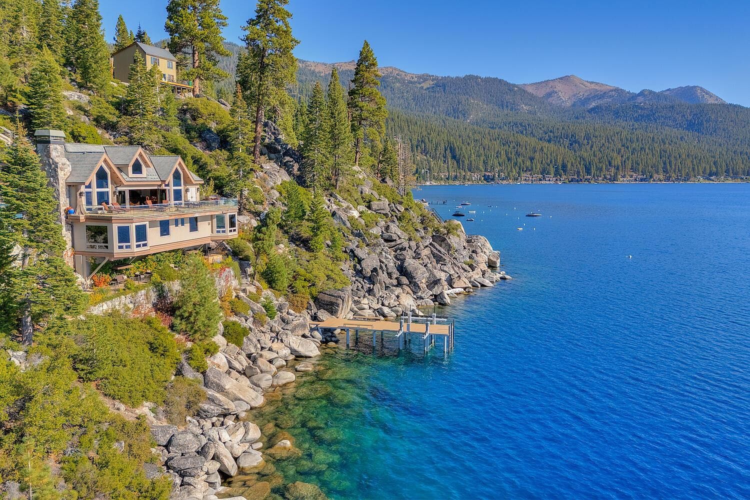This aerial view showcases a stunning waterfront property with a multi-level home perched on a rocky cliff overlooking a clear blue lake. The house features a deck, large windows, and a unique architectural design that blends seamlessly with the natural surroundings. A private dock extends into the water, enhancing the property's appeal for lake activities.