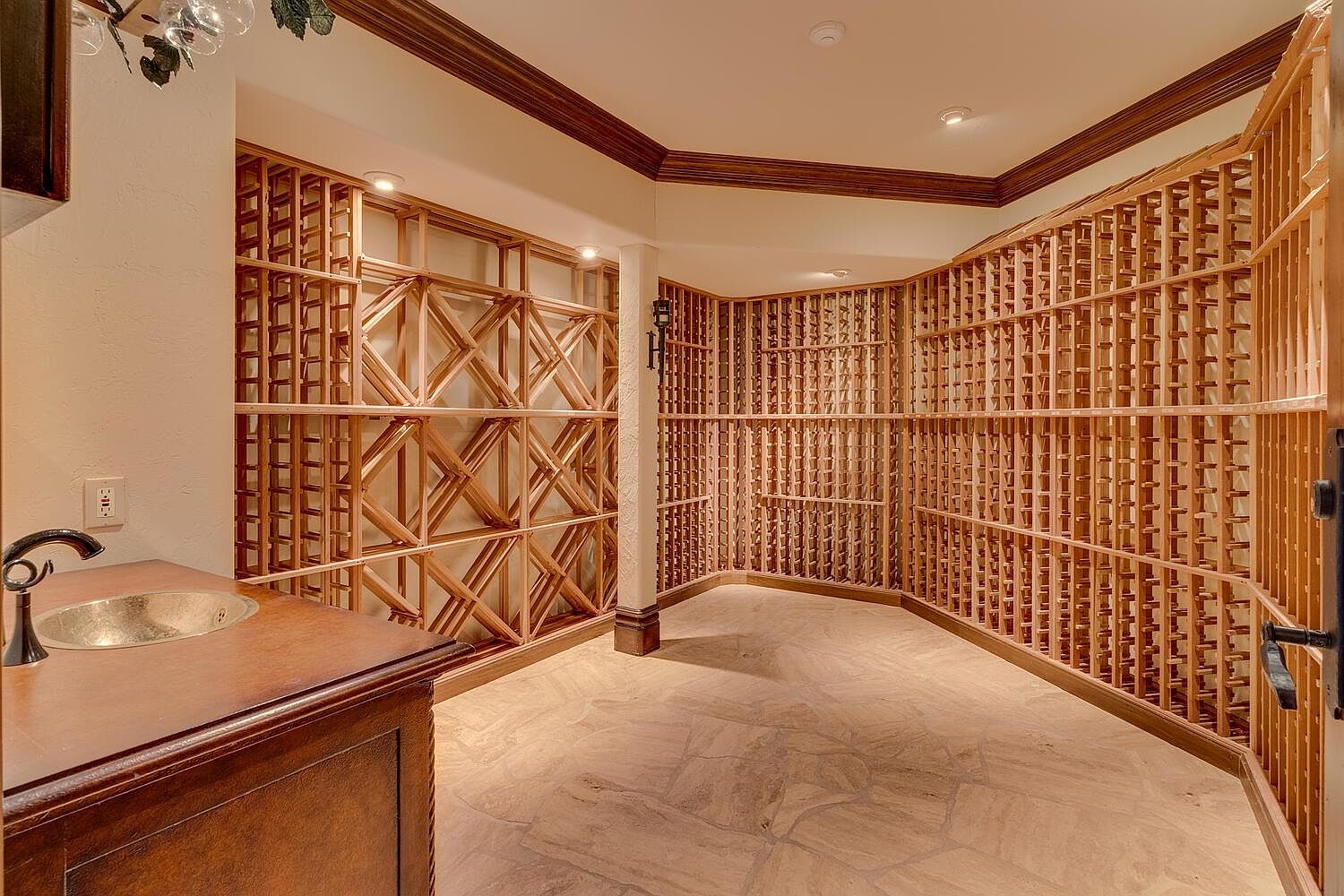 This is an interior shot of a well-appointed wine cellar. The room features extensive wooden wine racks covering the walls, a stone tile floor, and recessed lighting. A small sink and countertop area are visible in the foreground, suggesting a space for wine preparation and tasting.