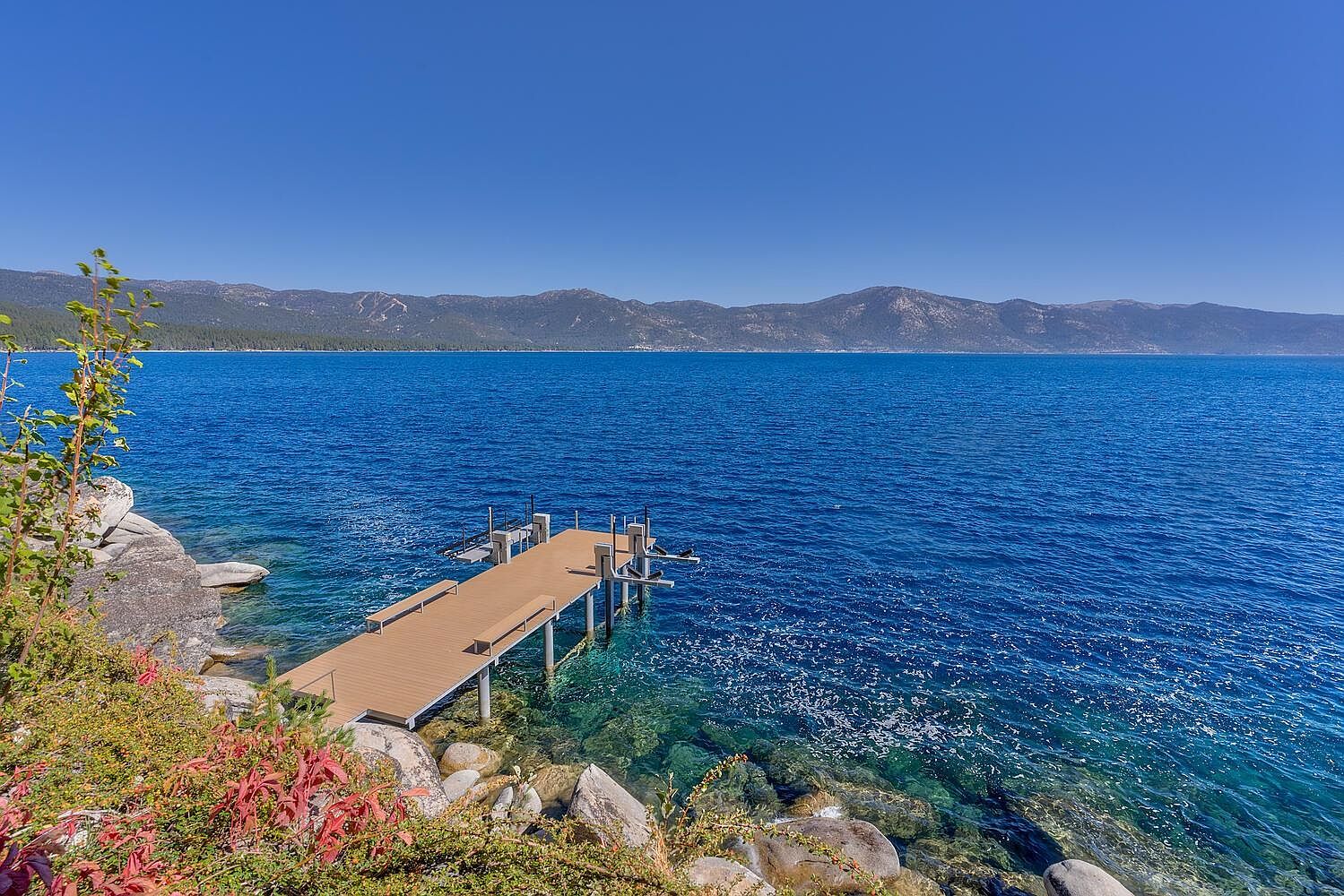 This image showcases a beautiful waterfront property featuring a private dock extending into a clear blue lake. The dock is constructed with a light brown material and includes benches. The surrounding landscape includes rocks and greenery, with mountains visible in the background under a clear blue sky, creating a serene and inviting atmosphere.