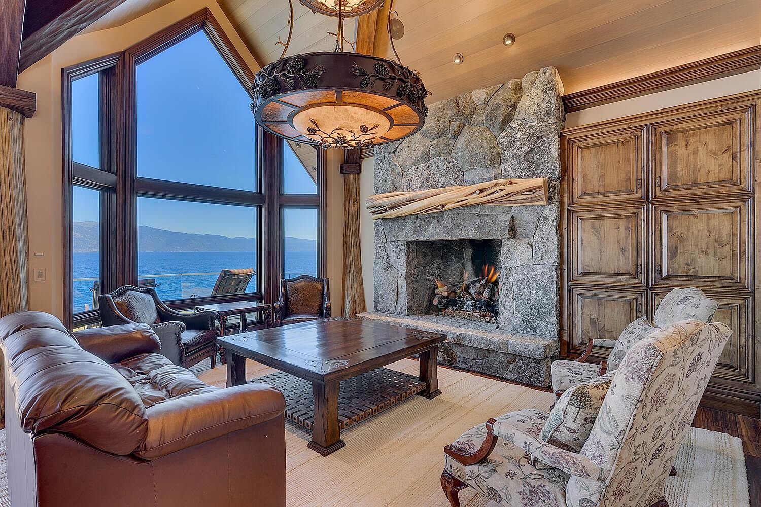 This is an interior shot of a luxurious living room featuring a stone fireplace with a wooden mantel, a large dark wood coffee table, and comfortable seating including a leather sofa and floral armchairs. Large windows offer a stunning view of the water, and an ornate chandelier hangs from the ceiling, creating a warm and inviting atmosphere.