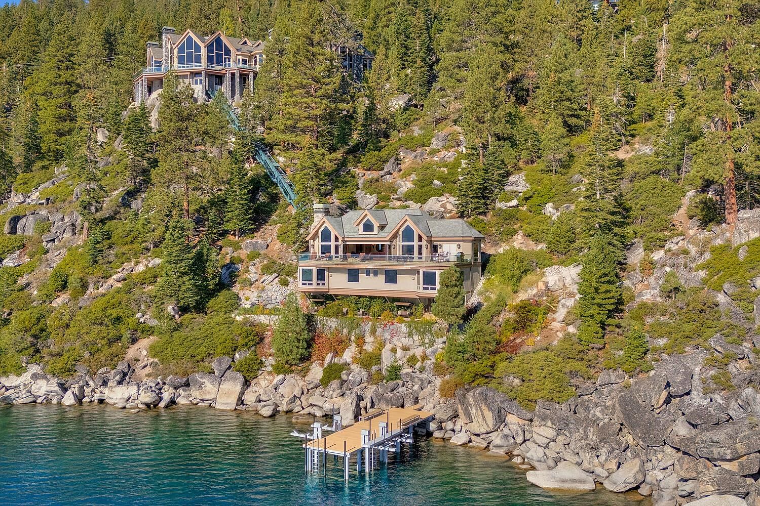 This aerial view showcases a stunning multi-level home nestled into a hillside overlooking a serene lake. The property features a private dock and a unique inclined elevator connecting the upper and lower levels. Lush greenery surrounds the home, enhancing its secluded and luxurious appeal.