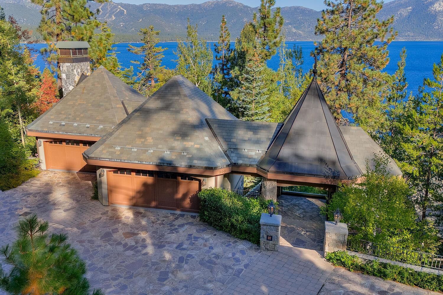 This aerial view showcases a luxurious home with unique architectural features, including multiple pyramidal roof structures and stone accents. The property boasts a spacious driveway, well-maintained landscaping, and a stunning backdrop of a serene lake and distant mountains. The overall impression is one of elegance and exclusivity.