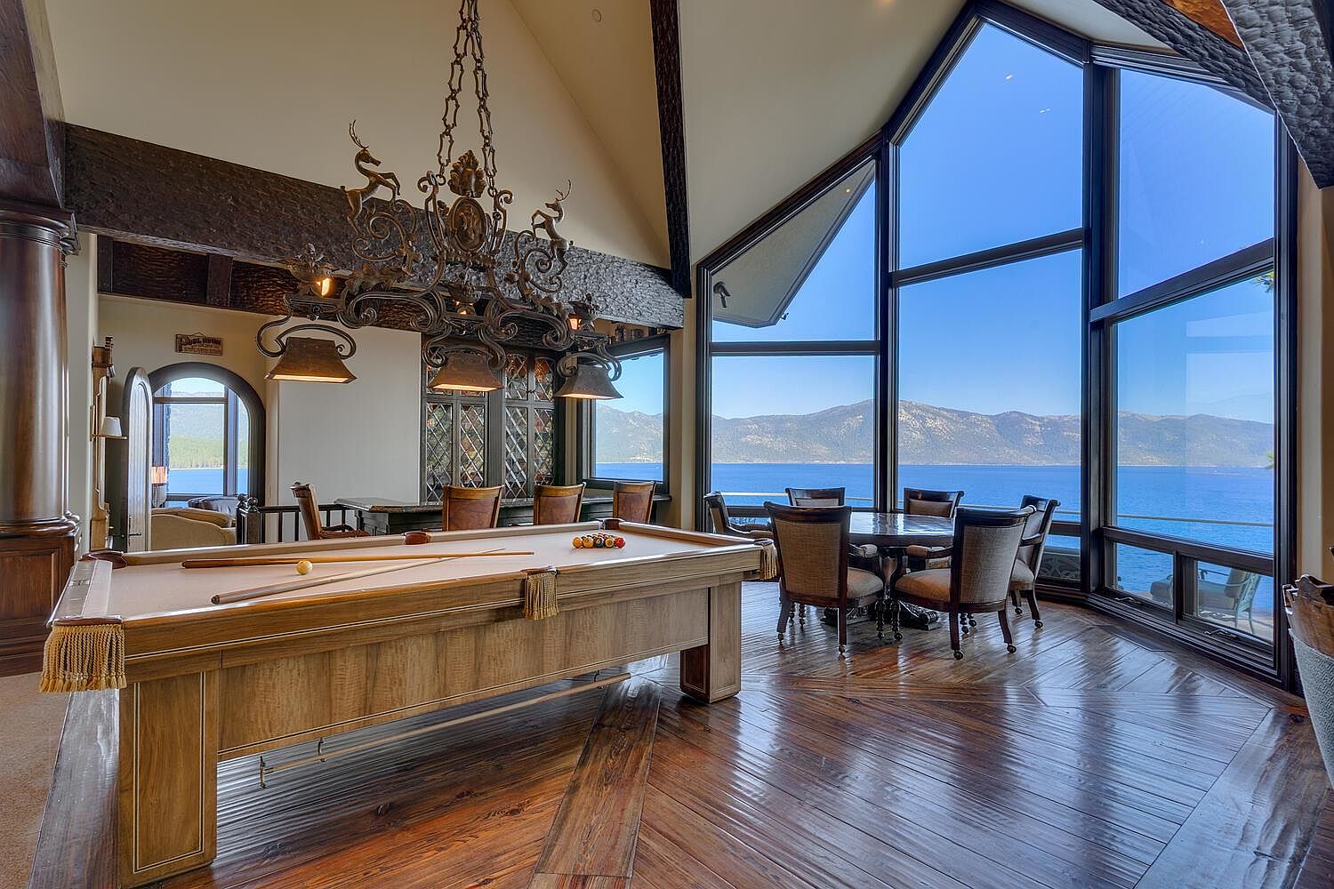 This is an interior shot of a luxurious living room featuring a pool table as a focal point. The room boasts floor-to-ceiling windows offering a stunning view of a lake and mountains, complemented by a large, ornate chandelier. The hardwood floors and elegant dining set contribute to the room's sophisticated ambiance.
