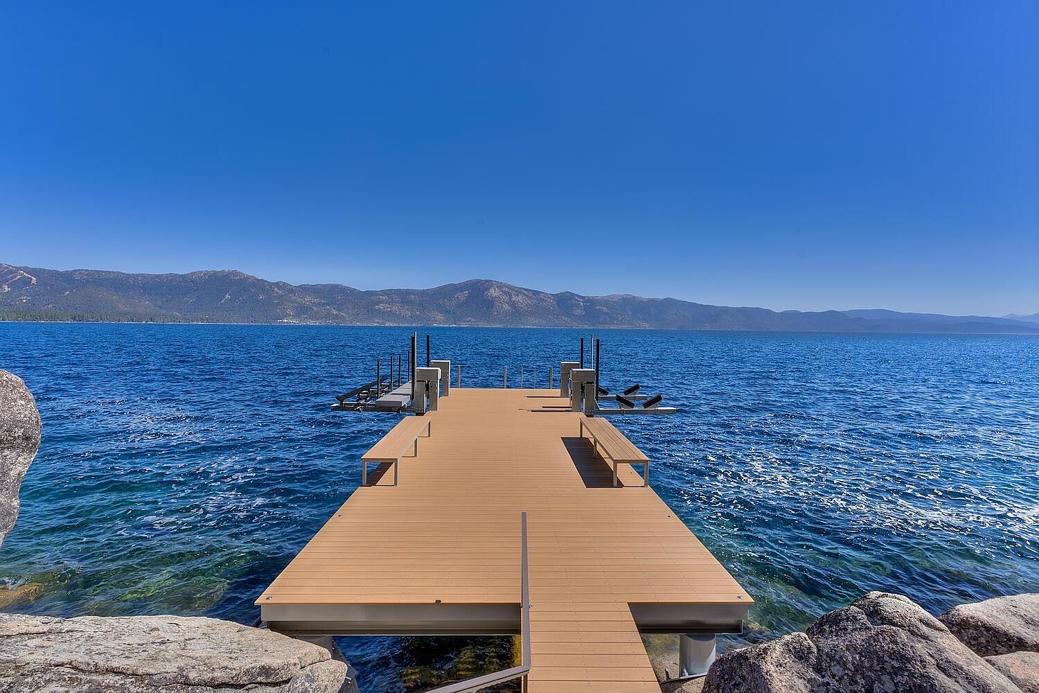 This image showcases a well-maintained wooden dock extending into a serene body of water, likely a lake, under a clear blue sky. The dock features built-in benches and boat lifts, suggesting a luxurious waterfront property. Mountains are visible in the background, adding to the scenic appeal and highlighting the property's desirable location.
