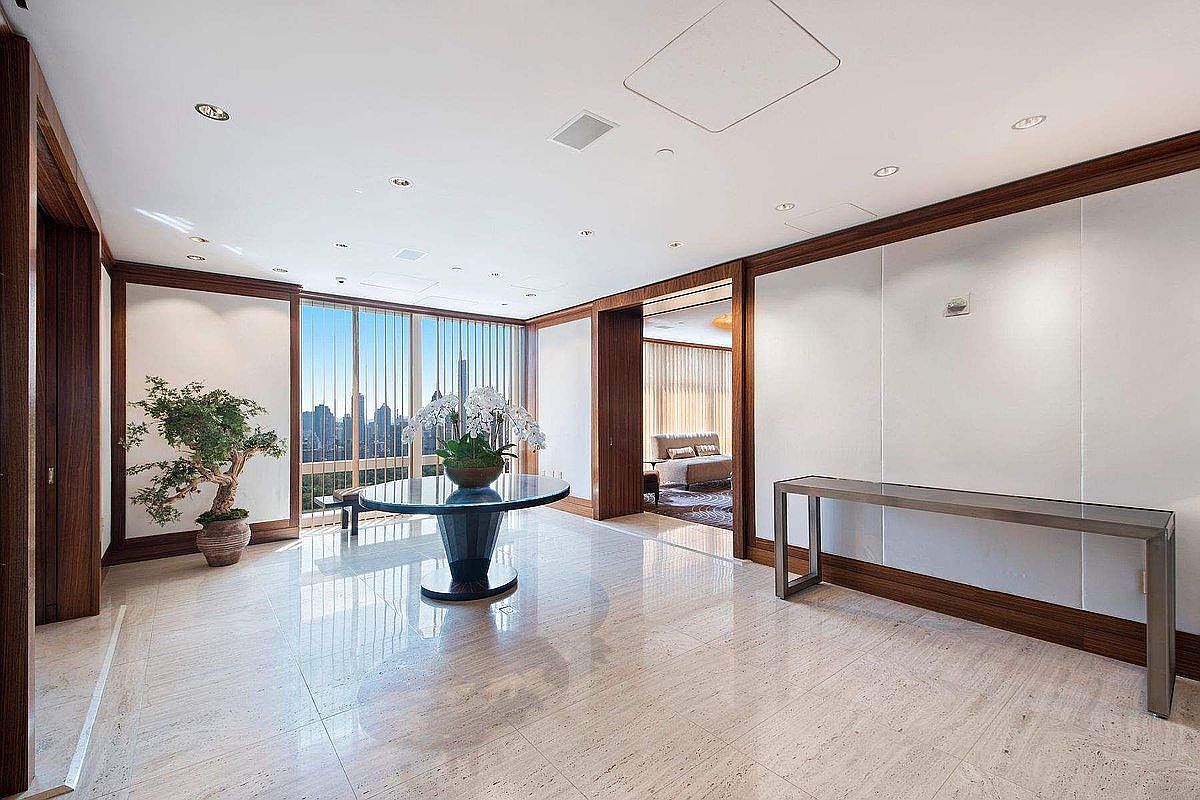 This elegant foyer or gallery space features polished light-colored stone flooring and rich wood trim that frames the walls and doorways. A central circular table with a floral arrangement serves as a focal point, while a sleek console table sits against the wall, all set against a backdrop of floor-to-ceiling windows offering a panoramic city view. The space exudes a sophisticated, high-end luxury aesthetic with clean lines and a bright, airy atmosphere.