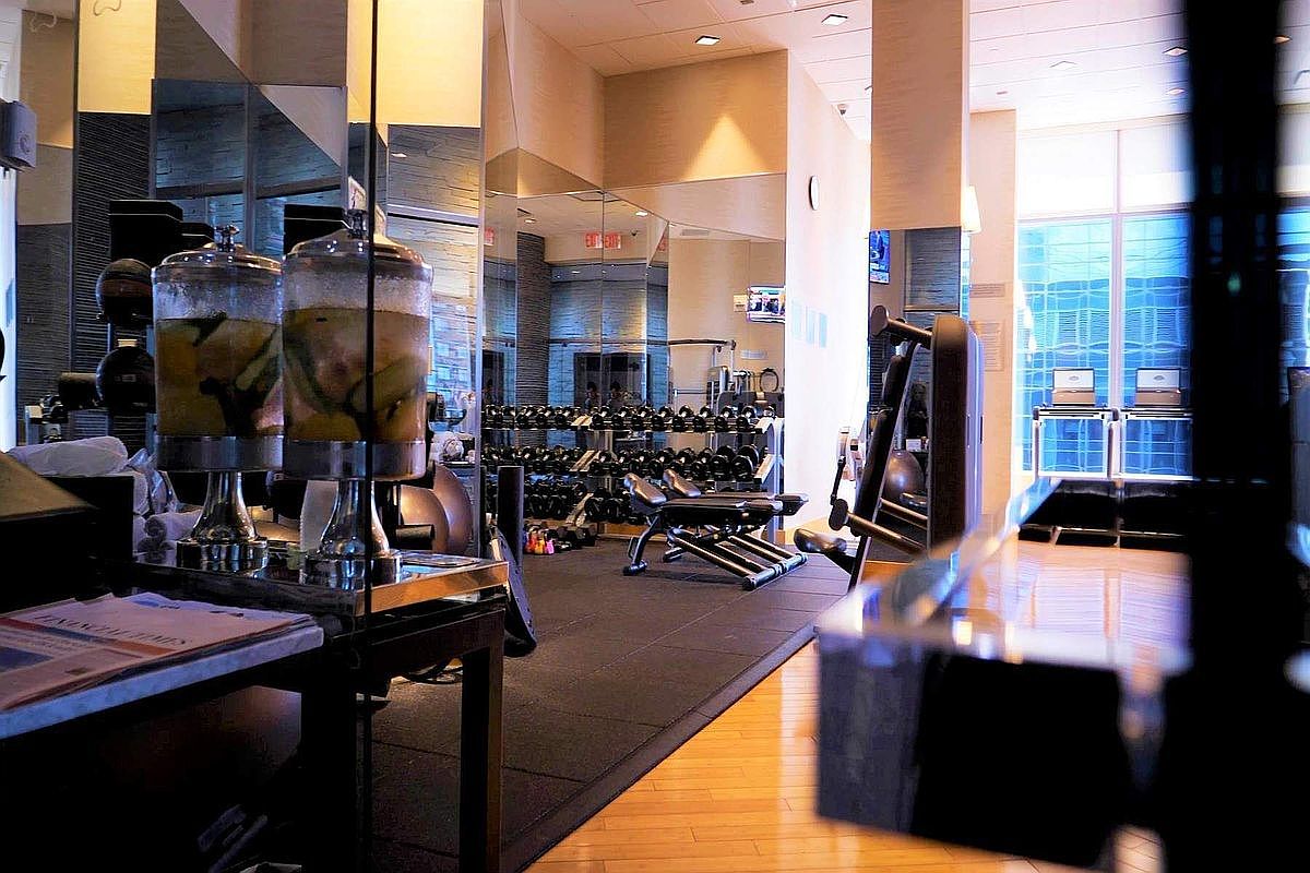 This well-appointed fitness center features a sleek, modern design with mirrored walls that enhance the sense of space and light. The room is equipped with a variety of professional-grade weight training equipment, including a rack of dumbbells and a weight bench, set against a backdrop of large windows that offer an expansive view. In the foreground, a refreshment station with infused water dispensers adds a touch of luxury, creating an inviting and high-end atmosphere for residents.