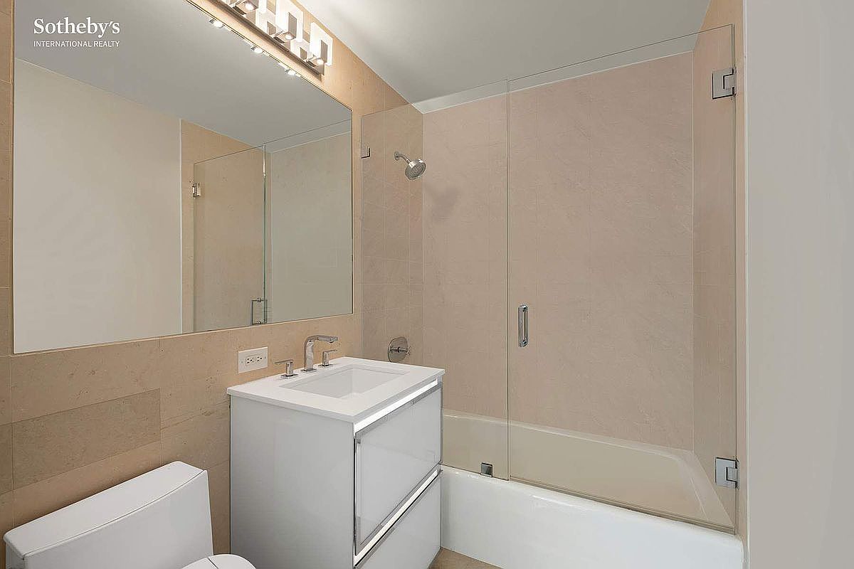 This clean and modern bathroom features a white vanity with a rectangular sink, a large wall-mounted mirror, and a shower-tub combination enclosed by a glass panel. The space is finished with neutral-toned wall tiles and a contemporary light fixture above the mirror. The perspective is a straight-on shot that highlights the efficient layout and bright, minimalist aesthetic of the room.