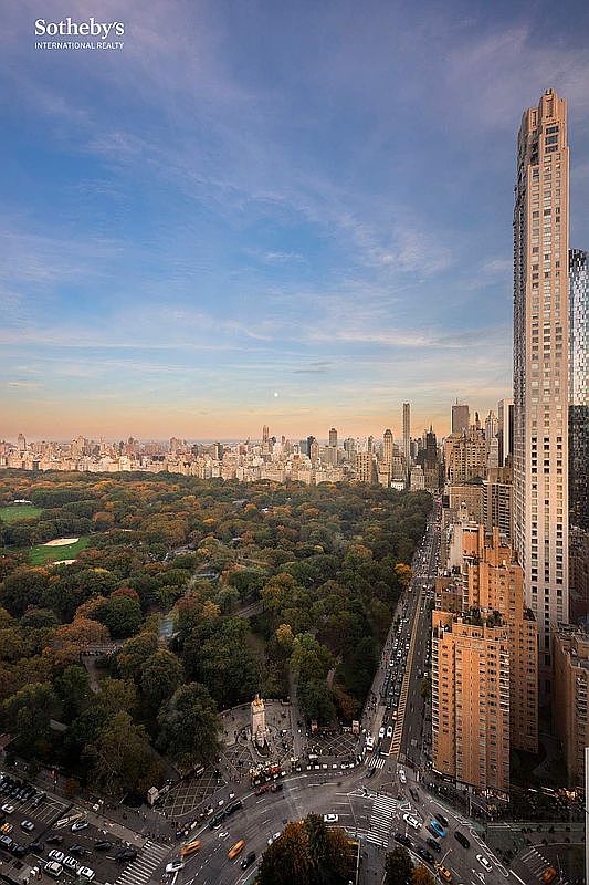 This high-angle aerial view captures a breathtaking perspective of Central Park in New York City, framed by the dense urban skyline. The image highlights the contrast between the lush, expansive greenery of the park and the towering, modern skyscrapers that line its perimeter, including a prominent, slender luxury residential tower in the foreground. The perspective provides a cinematic sense of scale, showcasing the vibrant city life and the iconic intersection below at dusk.