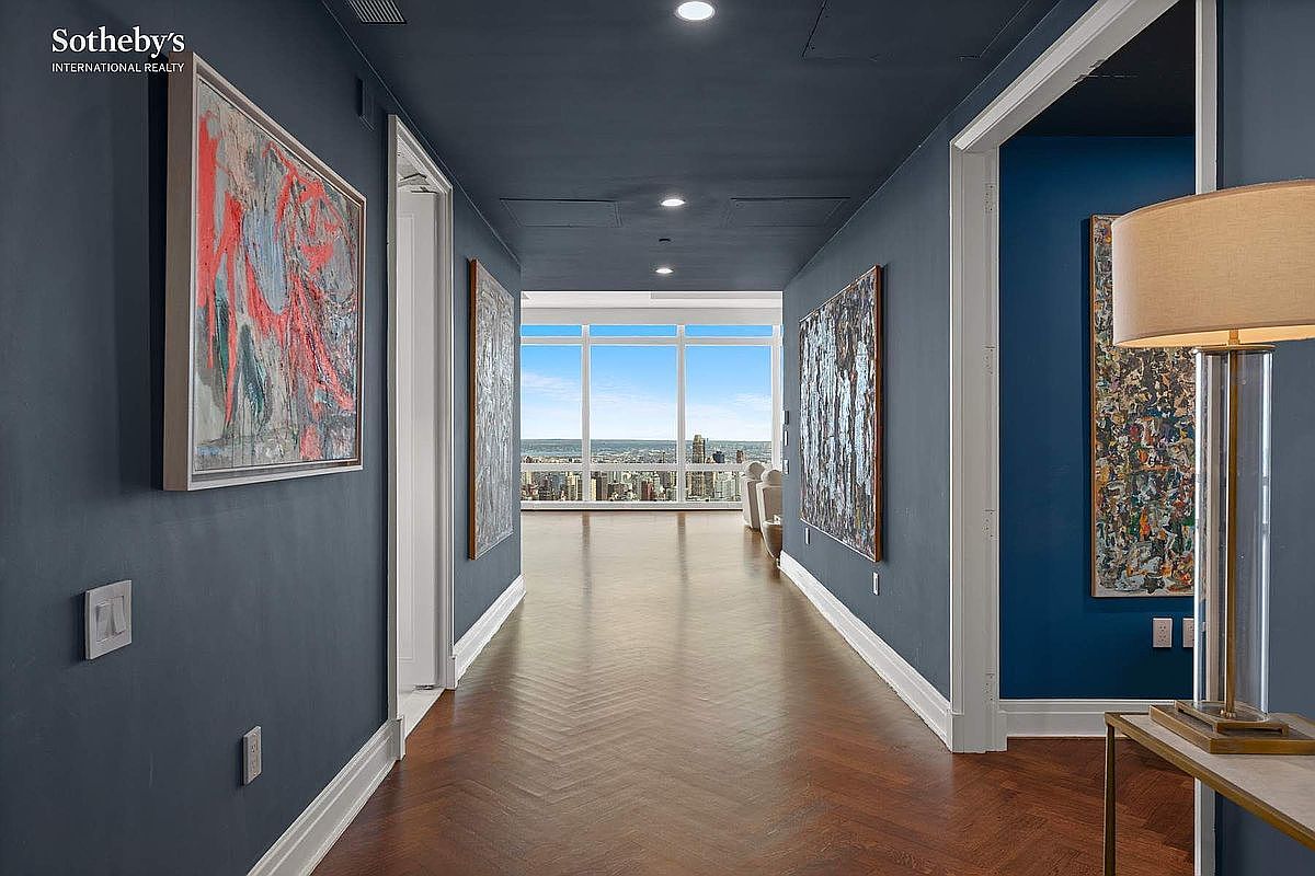This elegant hallway features deep navy blue walls adorned with large, vibrant abstract paintings, creating a sophisticated and artistic atmosphere. The space is anchored by rich, dark-stained herringbone hardwood floors that lead the eye toward a bright, floor-to-ceiling window offering expansive city views. A sleek table lamp in the foreground adds a warm, inviting glow, emphasizing the high-end, modern aesthetic of this luxury residence.