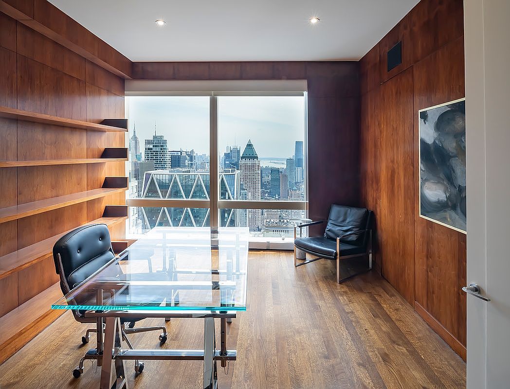 This sophisticated home office features rich, floor-to-ceiling wood paneling and a sleek glass-topped desk, creating a professional and modern atmosphere. A large window provides a breathtaking, high-rise panoramic view of the city skyline, serving as a dramatic focal point. The room is furnished with a black leather office chair and a matching accent chair, emphasizing a clean, minimalist aesthetic.