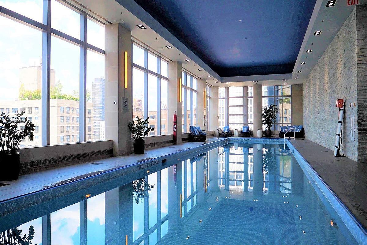This sophisticated indoor lap pool features floor-to-ceiling windows that offer expansive city views and bathe the space in natural light. The room is accented by a striking deep blue ceiling, sleek tiled flooring, and minimalist seating areas, creating a serene and luxurious atmosphere. The perspective captures the length of the pool, emphasizing its clean lines and modern architectural design.