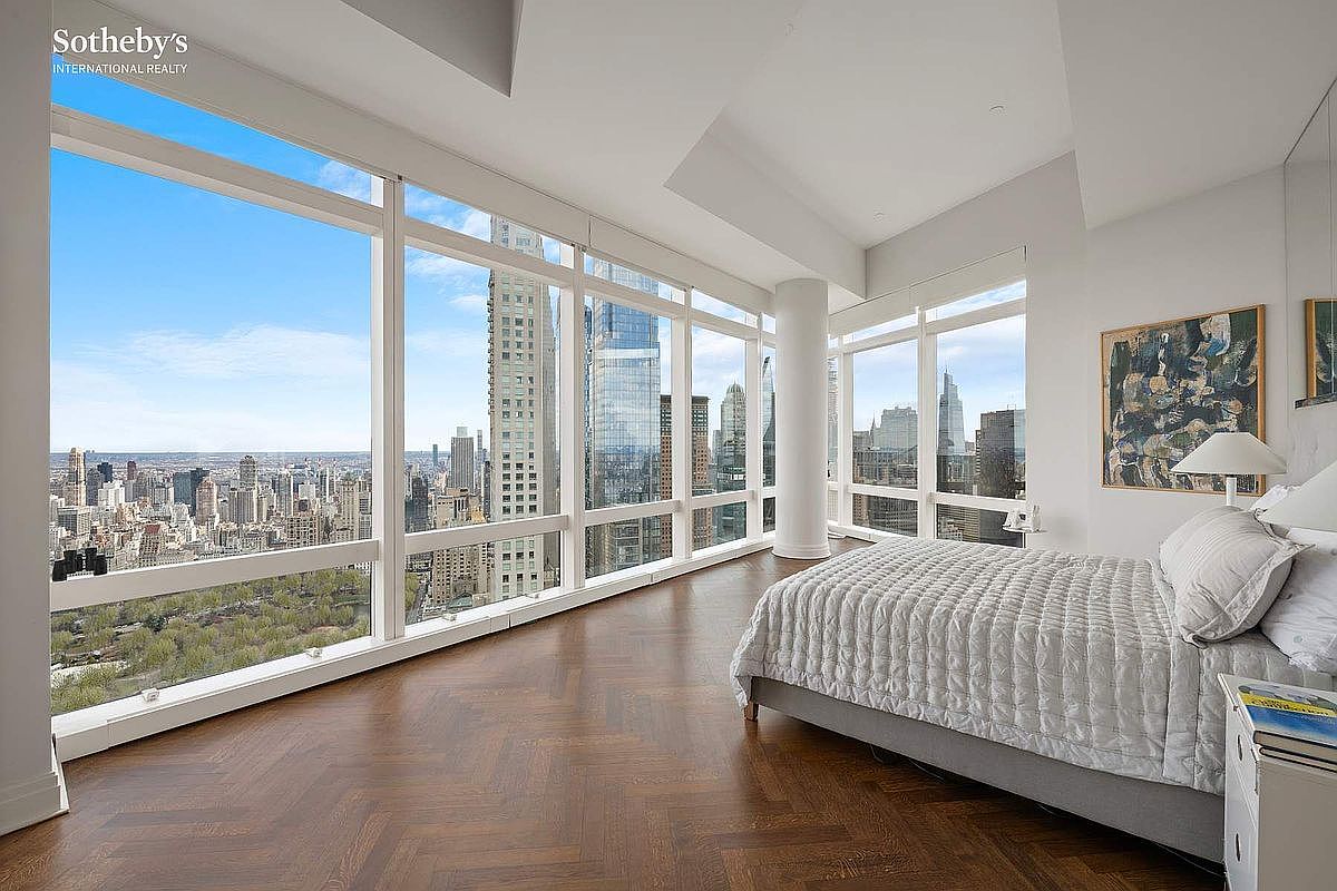 This luxurious primary bedroom features floor-to-ceiling windows that offer a breathtaking, panoramic view of the city skyline and Central Park. The room is anchored by a plush, light-colored bed with a quilted comforter and dark hardwood floors laid in a classic herringbone pattern. The minimalist, bright aesthetic emphasizes the expansive natural light and the high-end urban setting.