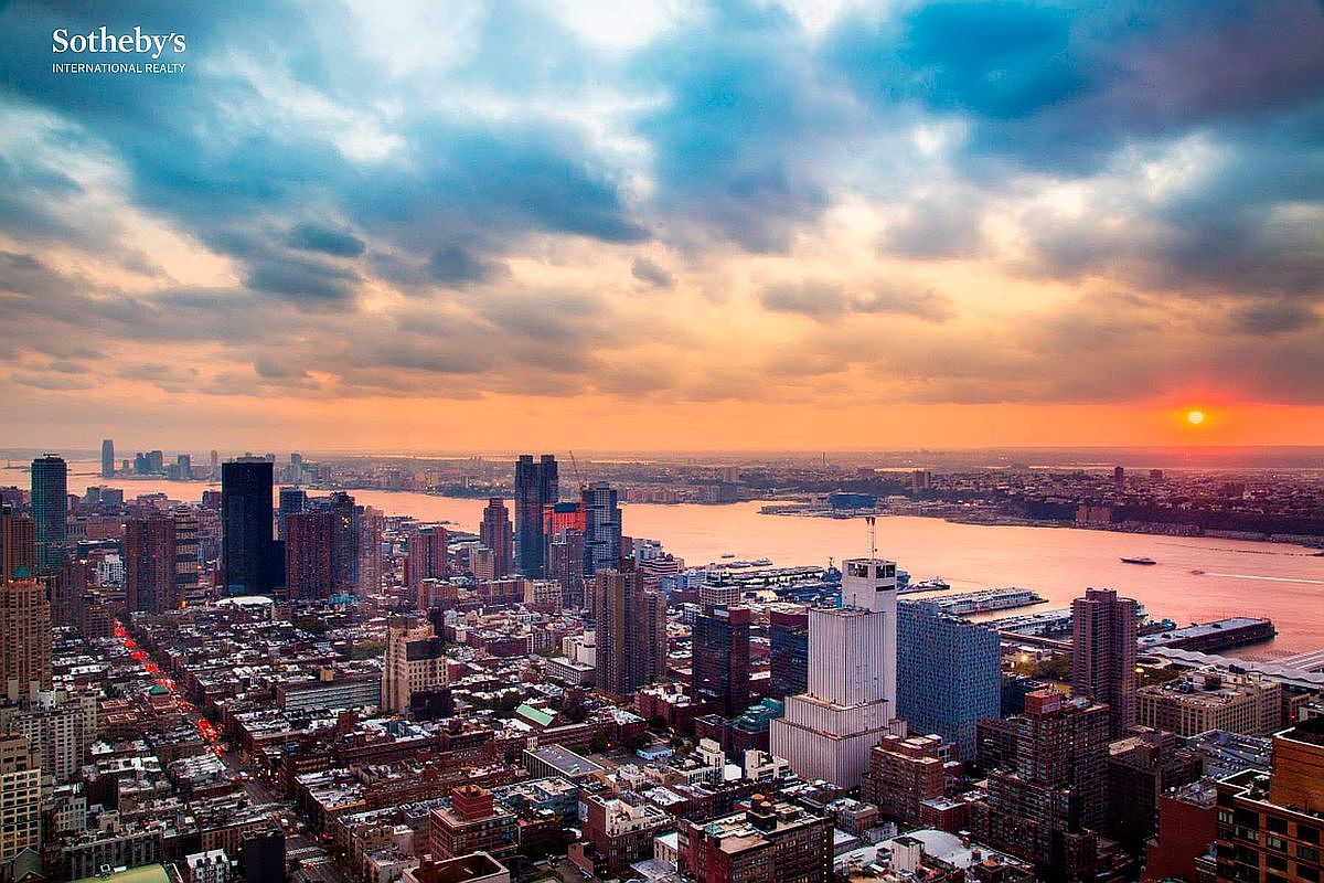 This stunning high-altitude aerial view captures a panoramic sunset over the New York City skyline, highlighting the dense urban architecture and the reflective waters of the Hudson River. The dramatic golden hour lighting casts a warm glow across the cityscape, emphasizing the scale and prestige of the location. The perspective provides a cinematic sense of grandeur, showcasing the vibrant metropolitan environment and the iconic waterfront.