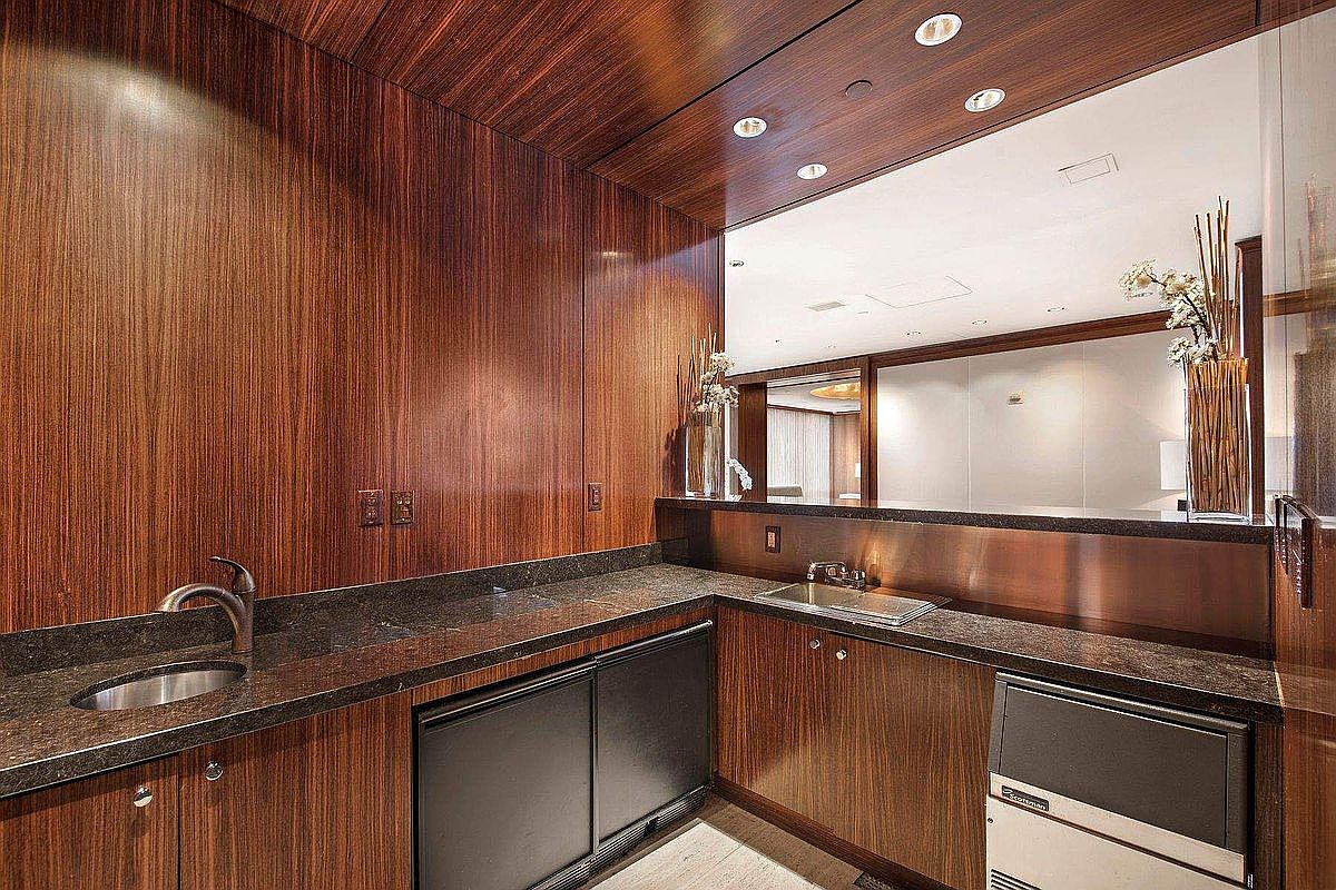 This sophisticated wet bar or kitchenette area features rich, dark wood paneling that extends from the walls to the ceiling, creating a warm and luxurious atmosphere. The space is equipped with dark granite countertops, two integrated sinks, and a built-in Scotsman ice maker, all accented by recessed lighting and a large mirror that reflects the adjacent room. The design is sleek and modern, offering a high-end, functional space perfect for entertaining.