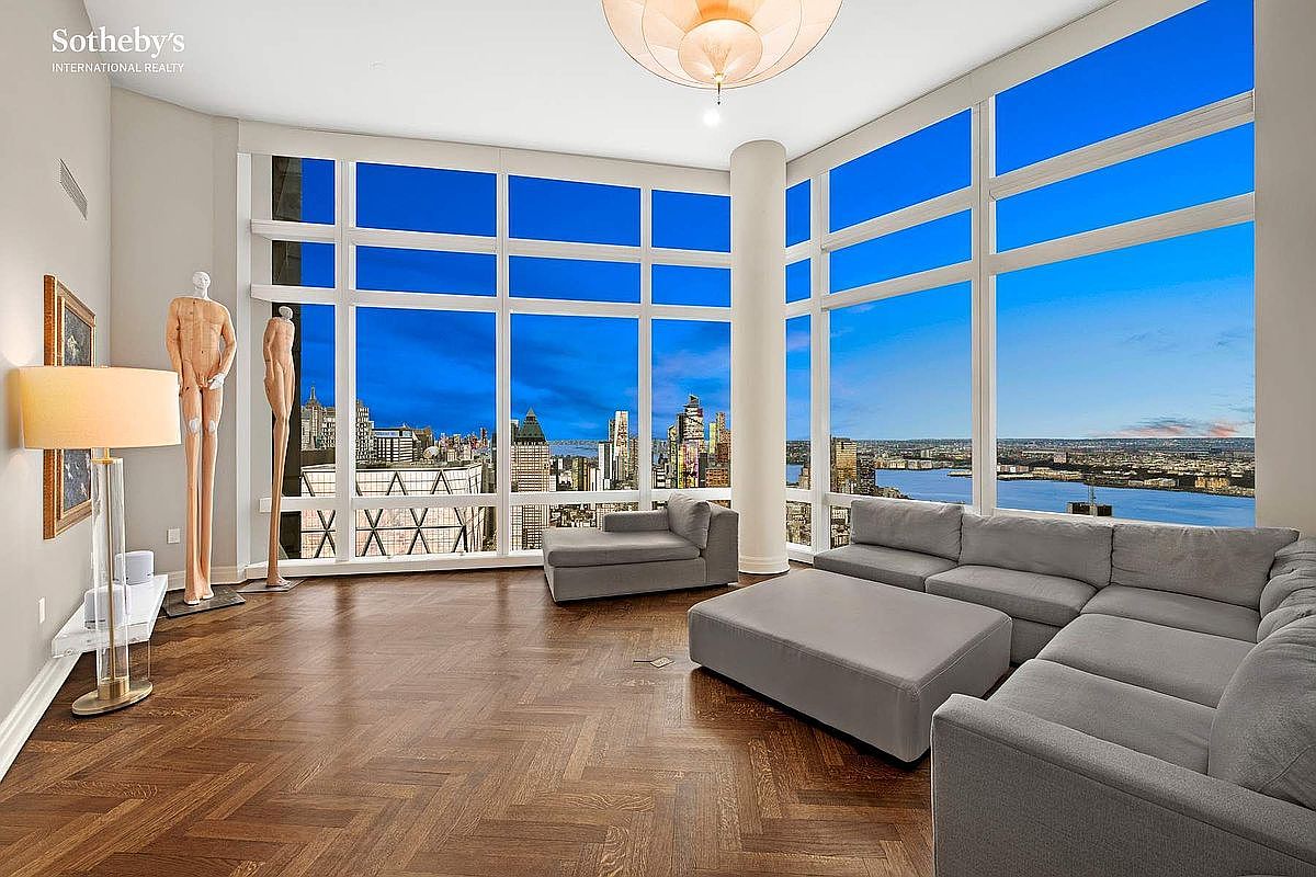 This expansive, high-ceilinged living room features floor-to-ceiling windows that offer a breathtaking panoramic view of the city skyline and river at dusk. The space is anchored by a large, plush grey sectional sofa and a matching ottoman, set against rich, dark herringbone-patterned hardwood floors. Two abstract human-form sculptures stand near the window, adding a unique artistic touch to the sophisticated, modern interior.