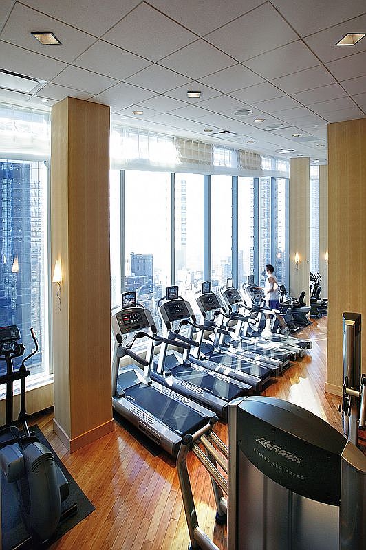This high-end fitness center features a row of professional-grade treadmills positioned against floor-to-ceiling windows that offer a stunning urban skyline view. The space is finished with warm hardwood flooring and light wood-paneled columns, creating a sophisticated and inviting atmosphere for exercise. A person is seen running on one of the treadmills, highlighting the functional and active nature of this luxury building amenity.