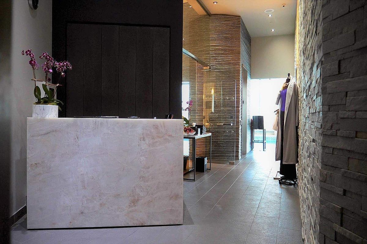 This sophisticated entryway features a striking, light-colored stone reception desk topped with a vibrant orchid arrangement, set against a dramatic dark accent wall. To the right, a textured stone wall leads the eye down a sleek hallway toward a glass-enclosed space, while a mannequin dressed in elegant attire adds a boutique-like touch. The perspective is eye-level, capturing a refined, modern aesthetic that blends luxury materials with clean, minimalist lines.