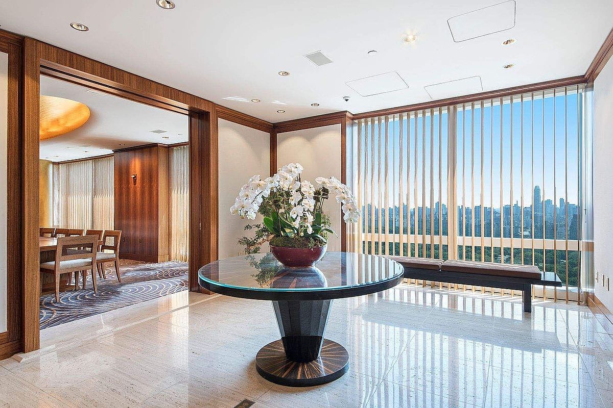 This elegant foyer features a polished marble floor and a striking circular glass-topped table as a central focal point, adorned with a large orchid arrangement. To the left, a wide doorway leads into a formal dining room with wood-paneled walls, while the right side showcases floor-to-ceiling windows with vertical blinds that offer a panoramic city view. The space exudes a sophisticated, high-end atmosphere with its clean lines, warm wood accents, and luxurious materials.