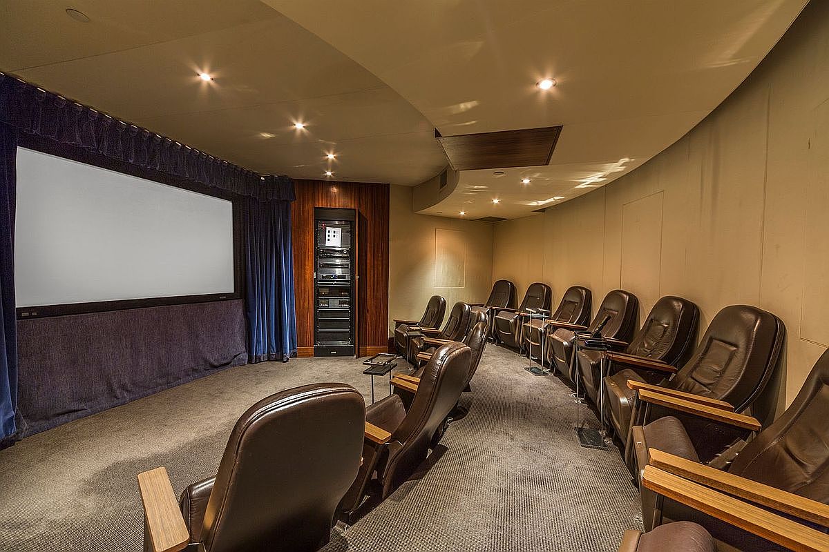 This image showcases a dedicated home theater room featuring tiered rows of plush, dark leather reclining seats facing a large projection screen. The room is designed with a curved ceiling, recessed lighting, and dark blue curtains, creating an immersive cinematic atmosphere. A tall equipment rack is built into the wood-paneled wall, emphasizing the high-end audio-visual capabilities of this private entertainment space.
