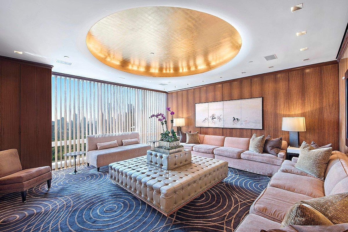 This luxurious living room features a sophisticated design with wood-paneled walls, a striking gold-leaf circular ceiling medallion, and a large, tufted ottoman centerpiece. The space is furnished with plush, light-toned sofas and a patterned blue carpet, all set against a backdrop of floor-to-ceiling windows that offer an expansive city view. The overall aesthetic is one of high-end elegance and refined comfort, perfect for upscale urban living.