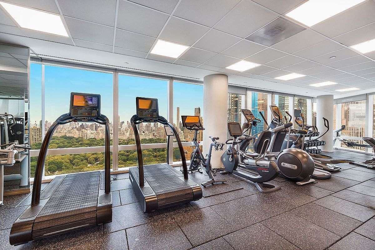 This high-end, modern home gym features a row of professional-grade treadmills, stationary bikes, and elliptical machines arranged neatly on a dark, durable rubber floor. Floor-to-ceiling windows provide an expansive, breathtaking panoramic view of a lush green park and the surrounding city skyline. The space is brightly lit by recessed ceiling panels, creating an inviting and energetic atmosphere for a workout.