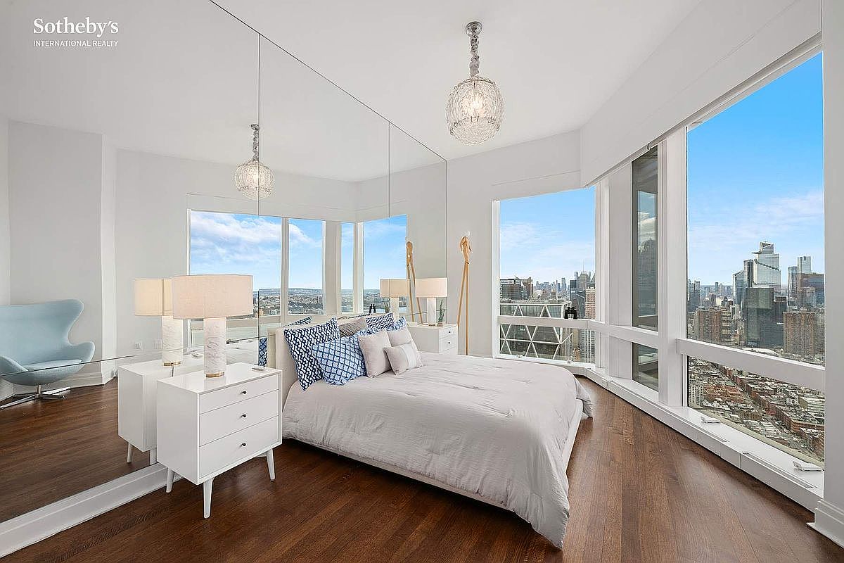 This sophisticated primary bedroom features floor-to-ceiling windows that offer a breathtaking, panoramic view of the city skyline. The room is elegantly appointed with a plush bed, mirrored wall accents that enhance the sense of space, and a modern, minimalist aesthetic. A stylish light blue accent chair and sleek white nightstands complete the luxurious, high-rise urban living experience.