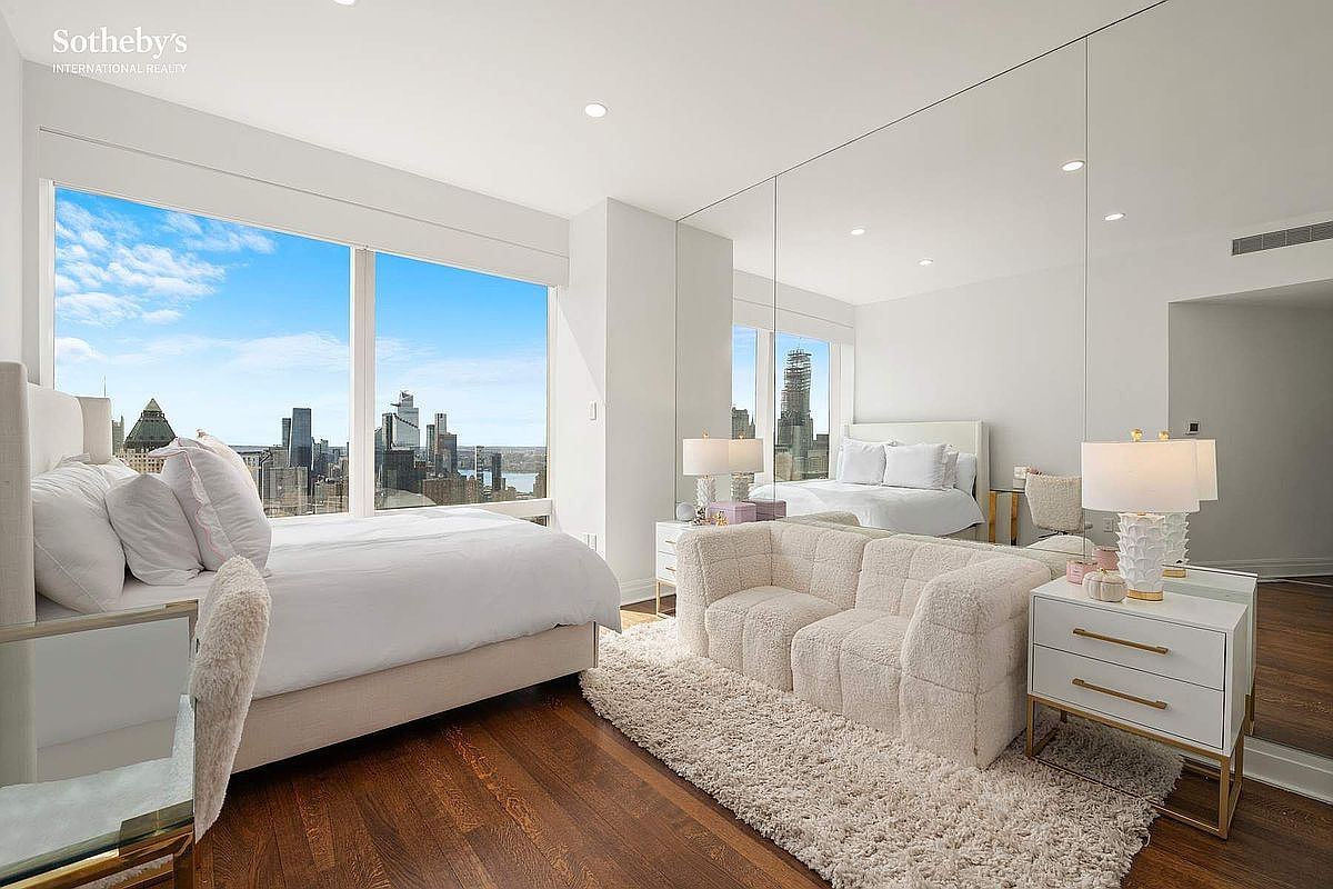 This luxurious primary bedroom features a plush, neutral-toned aesthetic with a large bed and a cozy, textured sofa set atop a soft area rug. A floor-to-ceiling mirrored wall enhances the sense of space, while expansive windows provide a breathtaking, unobstructed view of the city skyline. The room exudes a sophisticated, high-end urban atmosphere, perfectly blending comfort with modern architectural design.