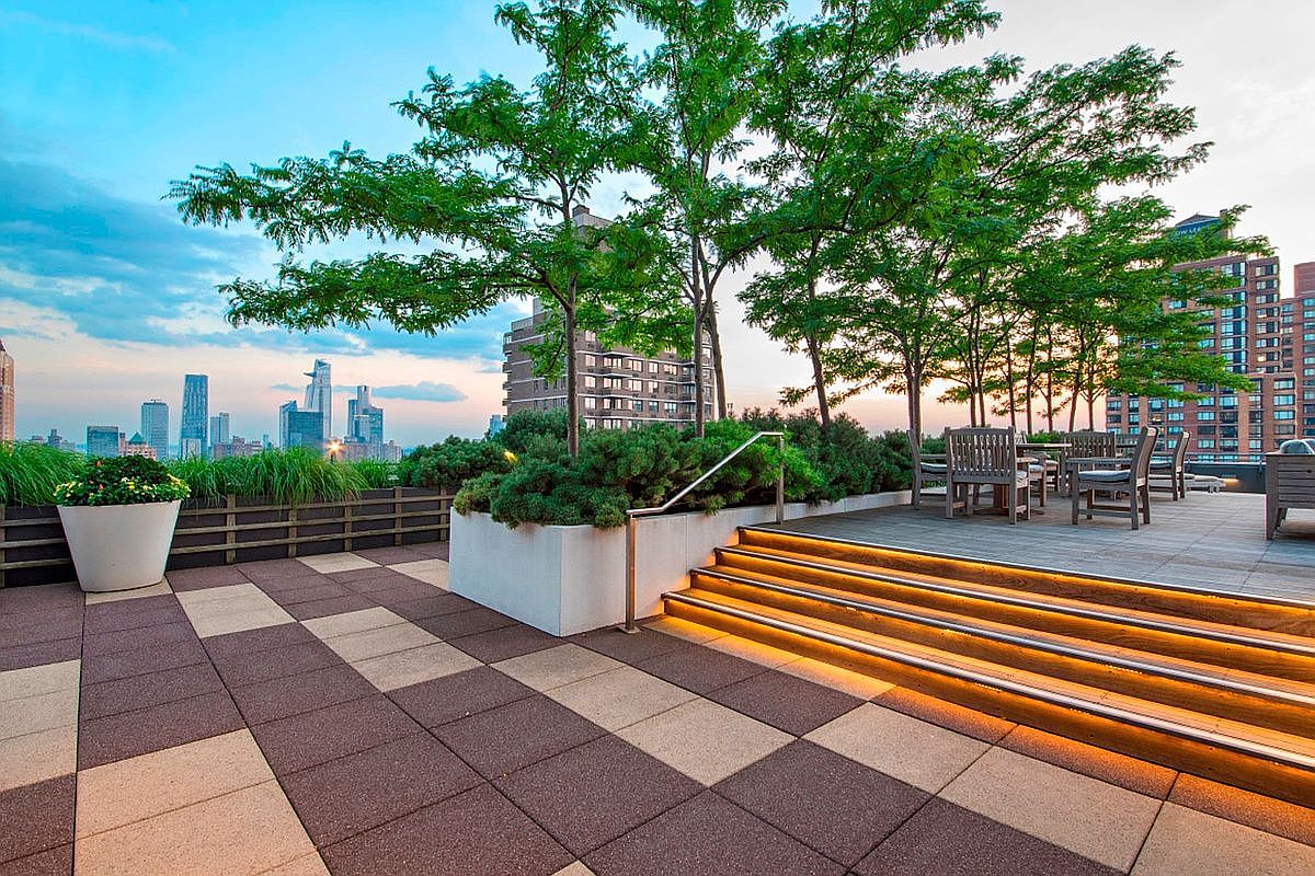 This sophisticated rooftop terrace offers a stunning urban oasis with a multi-level deck featuring integrated LED stair lighting and lush greenery. The space is furnished with wooden outdoor seating, providing a perfect vantage point to enjoy the panoramic city skyline at dusk. The combination of modern architectural design and natural elements creates an inviting and upscale atmosphere for outdoor relaxation.