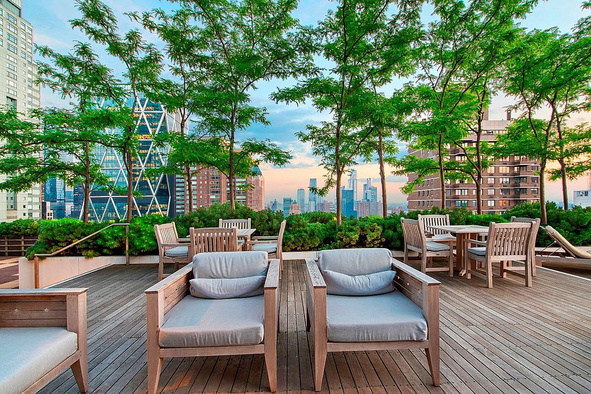 This expansive rooftop terrace features a warm wooden deck furnished with comfortable lounge chairs and dining sets, perfect for outdoor entertaining. Lush green hedges and mature trees frame the space, providing a natural contrast to the stunning, panoramic skyline view of the city's skyscrapers. The perspective is eye-level, capturing a serene and sophisticated urban oasis that feels both private and connected to the bustling city.