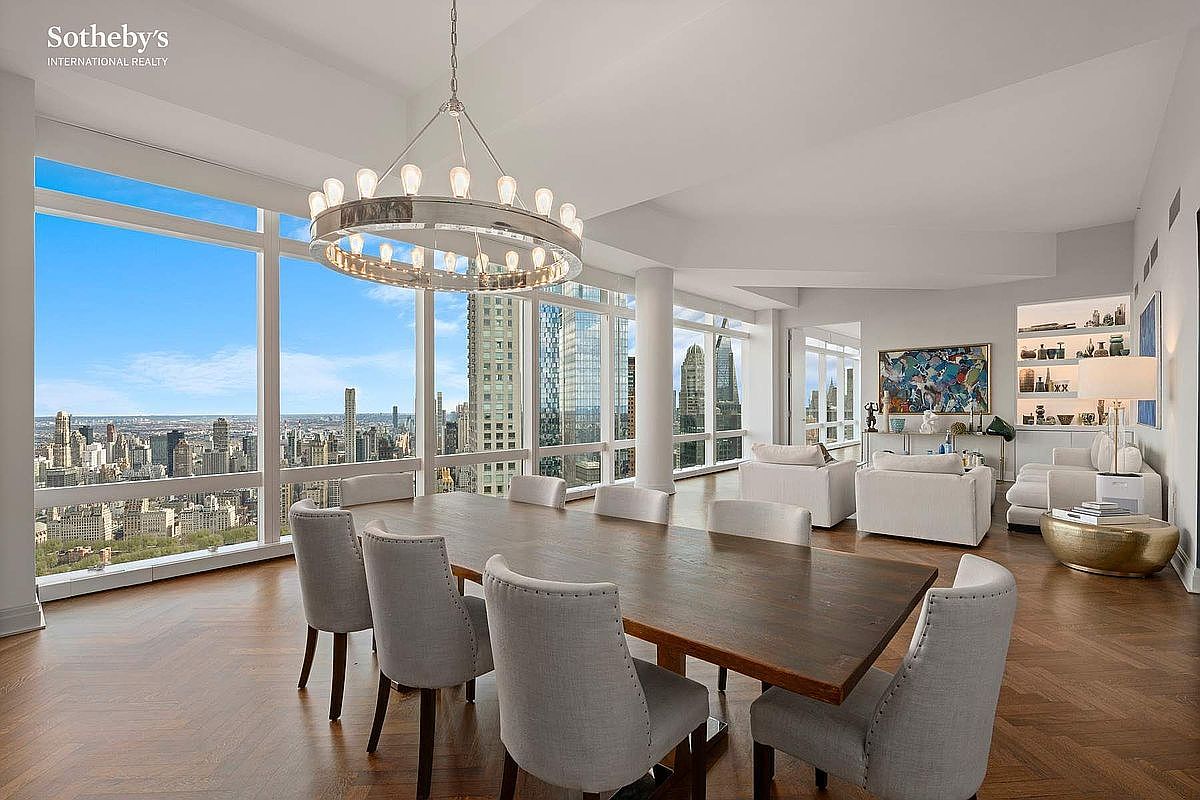 This sophisticated dining room features a large, dark wood table surrounded by eight upholstered chairs, positioned beneath a striking circular chandelier. The space is defined by floor-to-ceiling windows that offer a breathtaking, panoramic view of the city skyline, seamlessly blending indoor luxury with an urban backdrop. The open-concept layout flows into a bright living area, creating an airy and expansive atmosphere perfect for high-end entertaining.