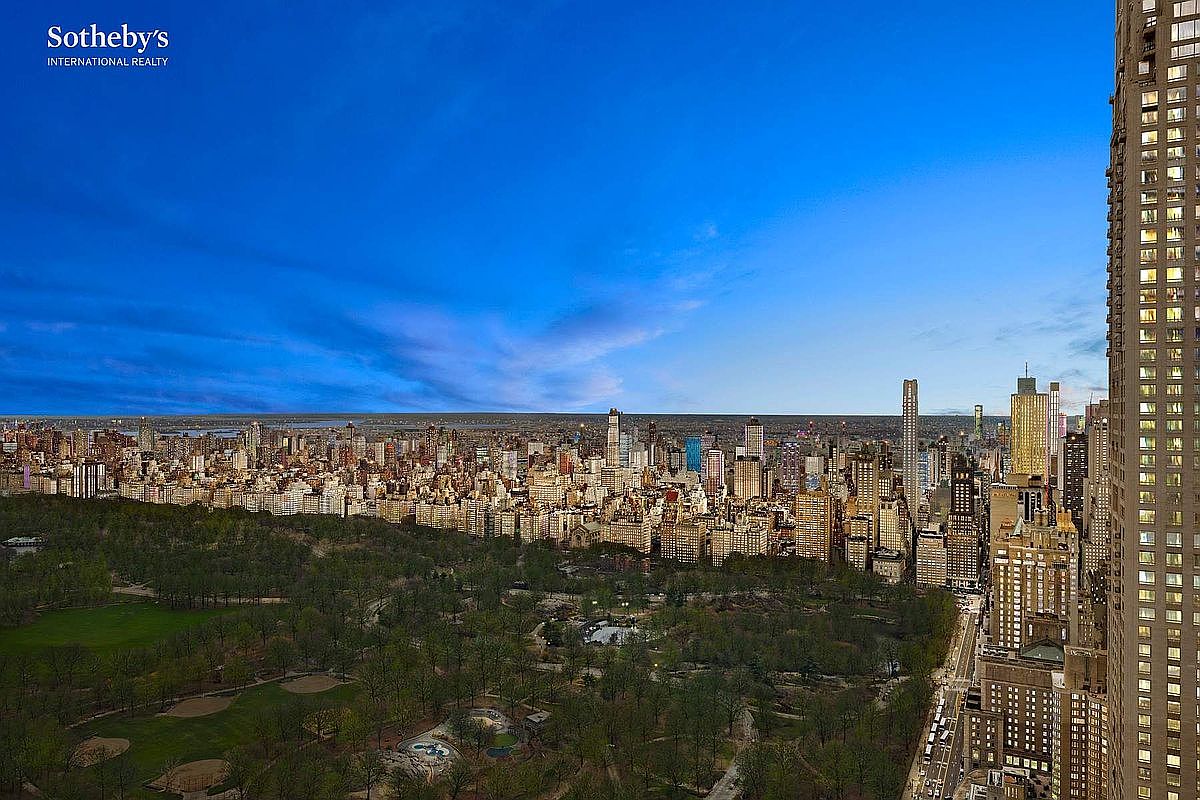 This high-altitude aerial view captures a breathtaking panoramic perspective of Central Park and the surrounding New York City skyline at dusk. The composition highlights the lush, expansive greenery of the park contrasted against the dense, illuminated urban architecture, emphasizing the property's prime location. The cinematic perspective provides a sense of grandeur and exclusivity, showcasing the iconic cityscape under a deep blue twilight sky.