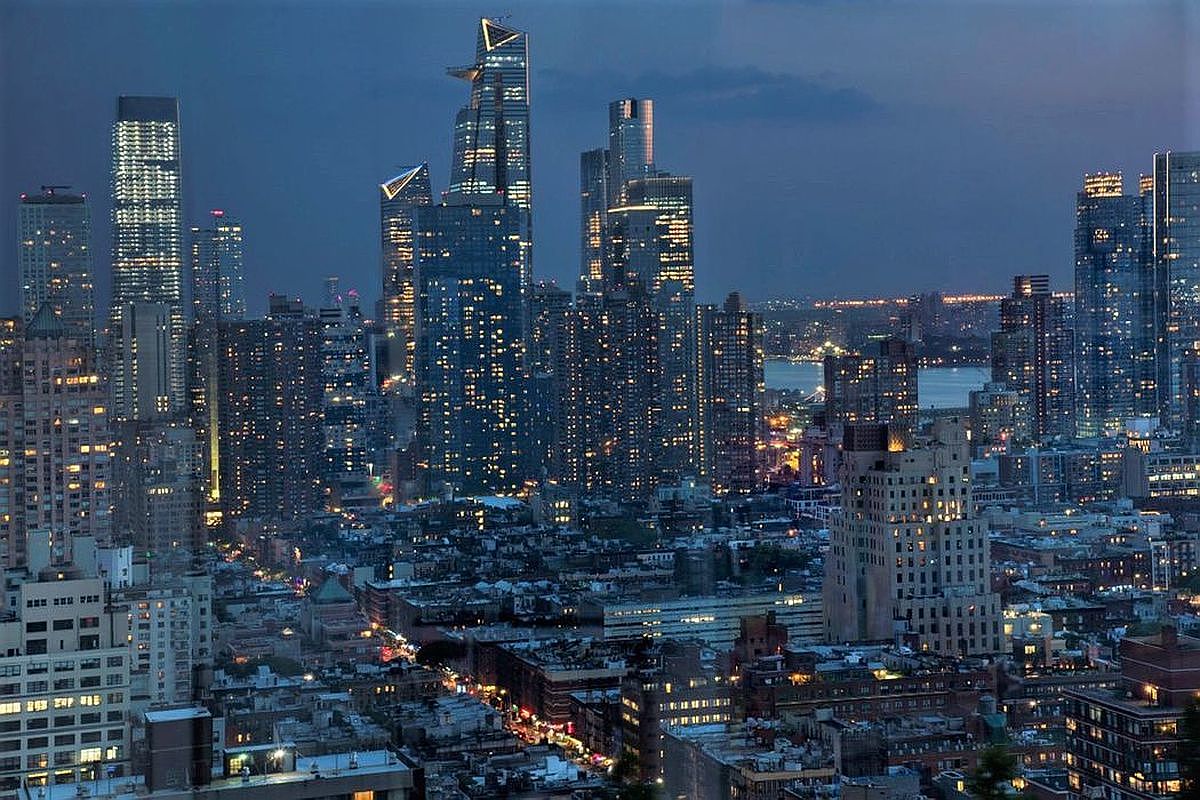 This stunning aerial view captures the dense, vibrant skyline of Manhattan at twilight, showcasing a mix of iconic modern skyscrapers and historic residential architecture. The scene is illuminated by the warm glow of thousands of apartment windows and streetlights, creating a sophisticated and energetic urban atmosphere. The perspective provides a sweeping, cinematic look at the city's vertical density and architectural diversity, perfect for highlighting a prime metropolitan location.