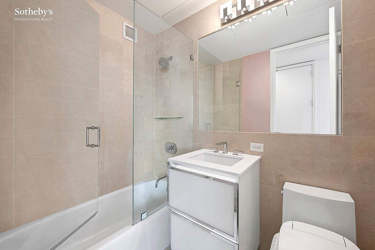 This modern, compact bathroom features a clean aesthetic with neutral-toned wall tiles and a sleek white vanity. The space includes a glass-enclosed shower-tub combination, a large wall-mounted mirror, and a contemporary toilet. The bright lighting and minimalist design create a functional and polished appearance suitable for a guest or secondary bathroom.