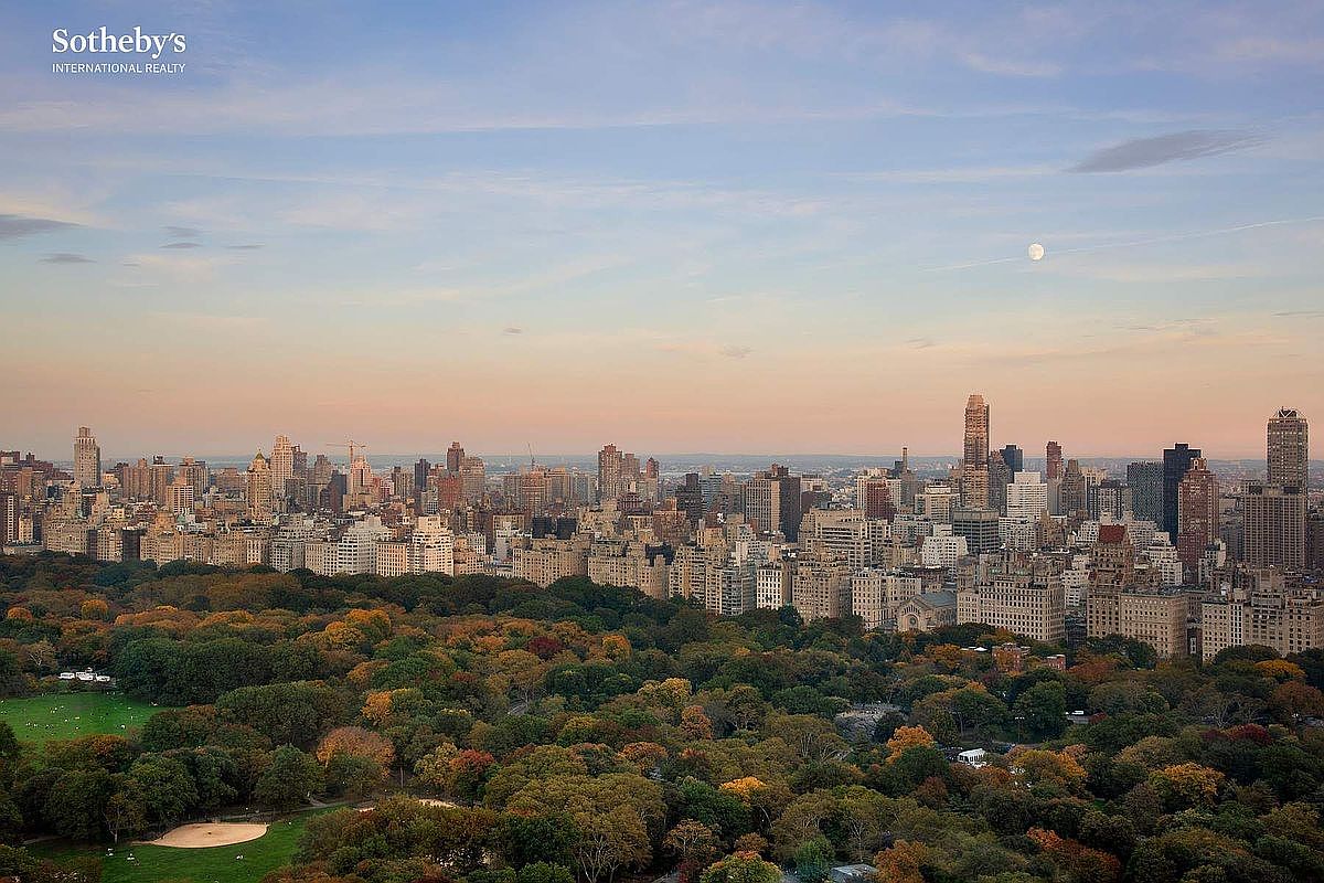 This high-angle aerial view captures a breathtaking panoramic vista of Central Park in New York City during the autumn season, with the dense, colorful canopy of trees transitioning into the iconic Manhattan skyline. The perspective emphasizes the property's prime location, showcasing the contrast between the lush greenery of the park and the dense urban architecture under a soft, twilight sky featuring a visible moon. The image conveys a sense of luxury, exclusivity, and the quintessential New York City lifestyle.
