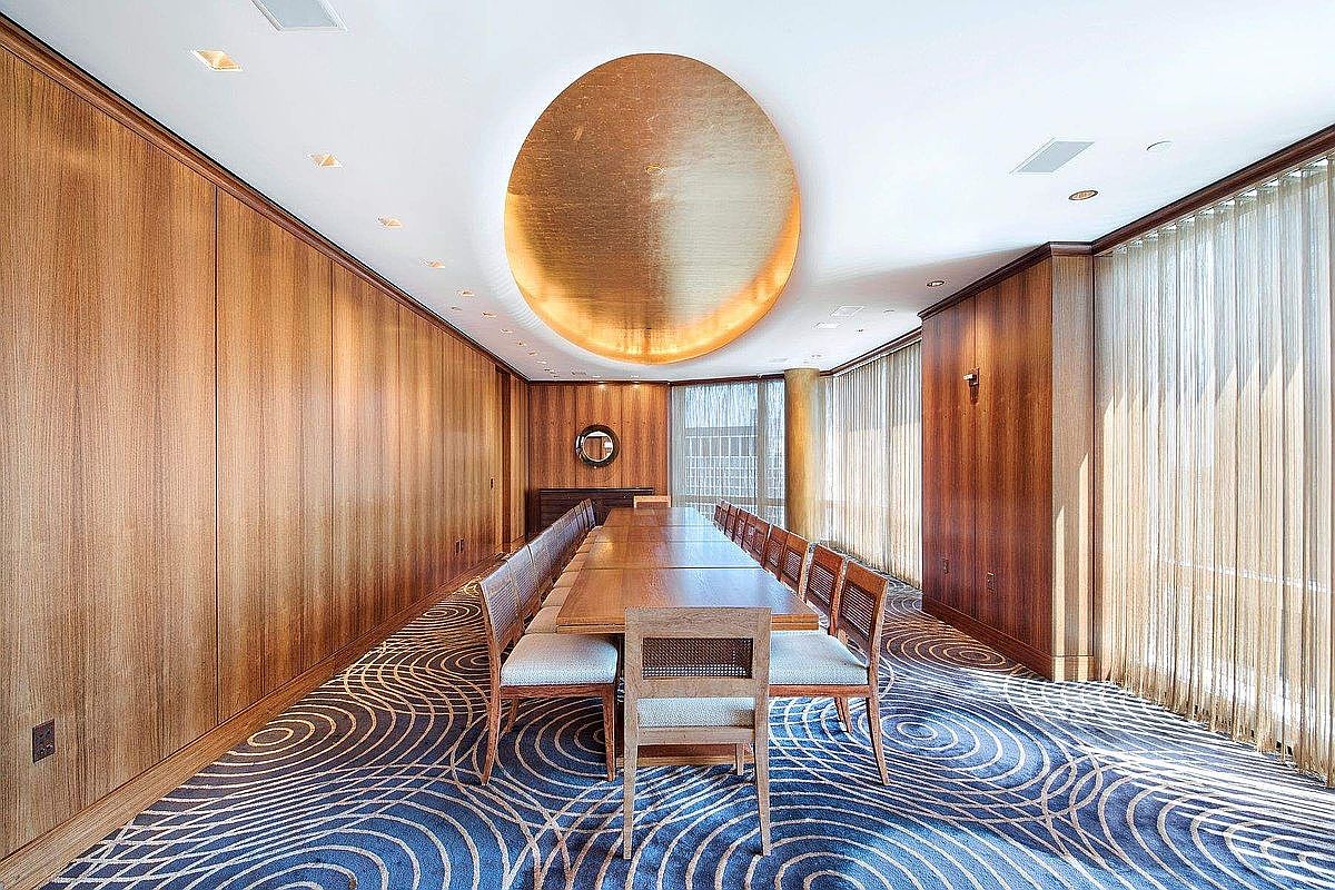 This sophisticated formal dining room features extensive wood-paneled walls and a striking, large circular gold-leaf ceiling fixture that serves as a dramatic focal point. A long, rectangular wooden table is surrounded by matching chairs with light-colored upholstery, set against a bold, patterned blue and gold carpet. The room is framed by floor-to-ceiling windows draped in sheer curtains, creating an elegant, high-end atmosphere suitable for professional meetings or formal gatherings.