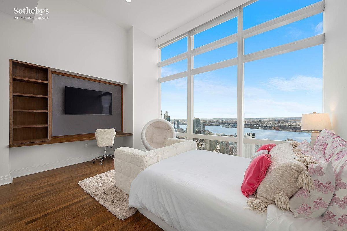 This bright and airy bedroom features floor-to-ceiling windows that offer a stunning, expansive view of the river and city skyline. The room is furnished with a plush white bed, a unique circular accent chair, and a built-in wooden media console with integrated shelving. The combination of warm hardwood floors and the dramatic natural light creates a sophisticated and serene atmosphere.