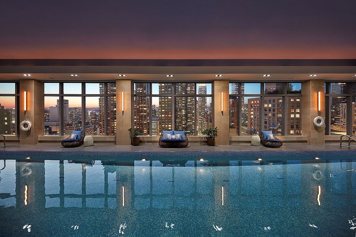 This luxurious indoor swimming pool area features a stunning panoramic view of a city skyline at dusk through floor-to-ceiling windows. The space is elegantly appointed with wicker lounge chairs, blue cushions, and potted plants, creating a serene and sophisticated atmosphere. The calm, reflective water of the pool emphasizes the high-end, tranquil nature of this urban amenity.