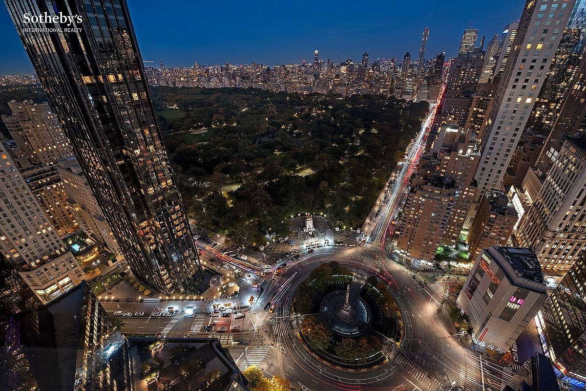 This stunning high-altitude aerial view captures the iconic Columbus Circle in New York City at twilight, showcasing the vibrant intersection of urban architecture and the lush greenery of Central Park. The perspective highlights the dramatic contrast between the illuminated skyscrapers and the dark, expansive parkland, emphasizing the prime location of the surrounding luxury real estate. The long-exposure light trails from traffic create a dynamic, cinematic sense of movement through the heart of the city.