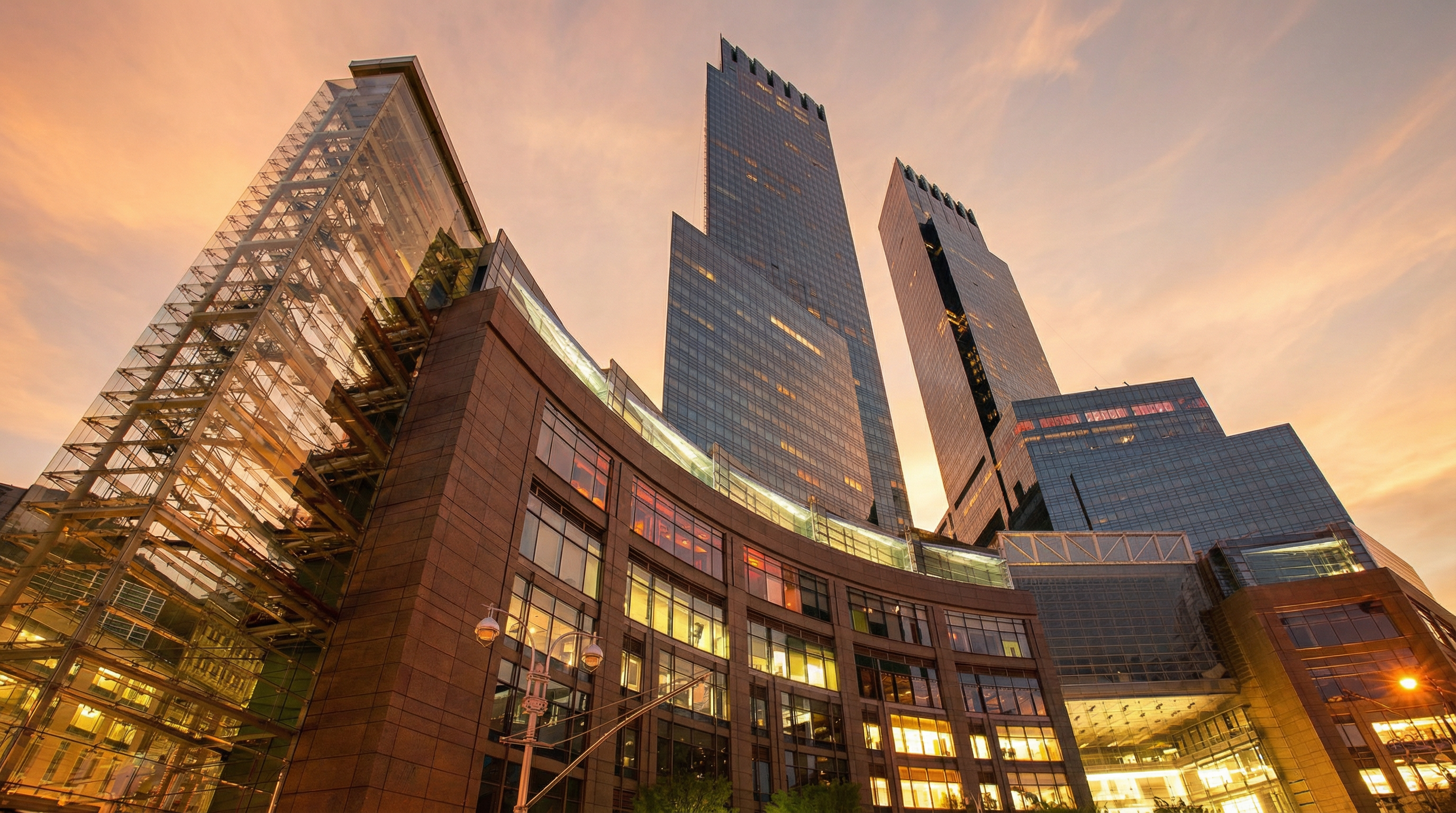 This low-angle, cinematic shot captures the imposing scale of a modern skyscraper complex at dusk, featuring a striking contrast between the warm, illuminated lower levels and the cool, blue-toned glass towers reaching into the twilight sky. The architecture showcases a blend of contemporary glass curtain walls and a curved, multi-story podium with intricate structural steel elements. The perspective emphasizes the height and urban sophistication of the development, creating a sense of grandeur and metropolitan luxury.