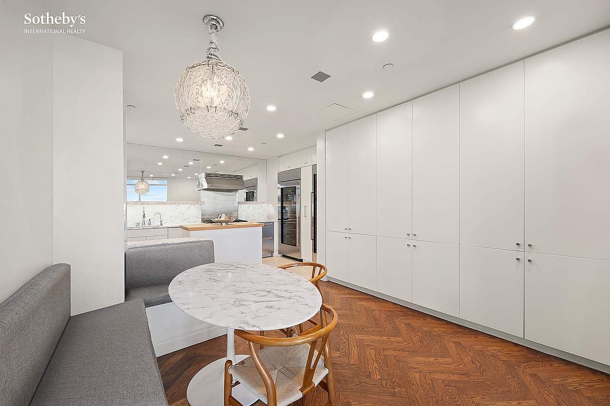 This modern kitchen features a sleek, minimalist design with floor-to-ceiling white cabinetry and rich herringbone-patterned hardwood floors. A marble-topped bistro table with wooden chairs and a built-in banquette creates a cozy dining nook, while a unique, textured spherical pendant light serves as a focal point. The space seamlessly transitions into a functional kitchen area with stainless steel appliances and reflective surfaces, offering a sophisticated and airy atmosphere.