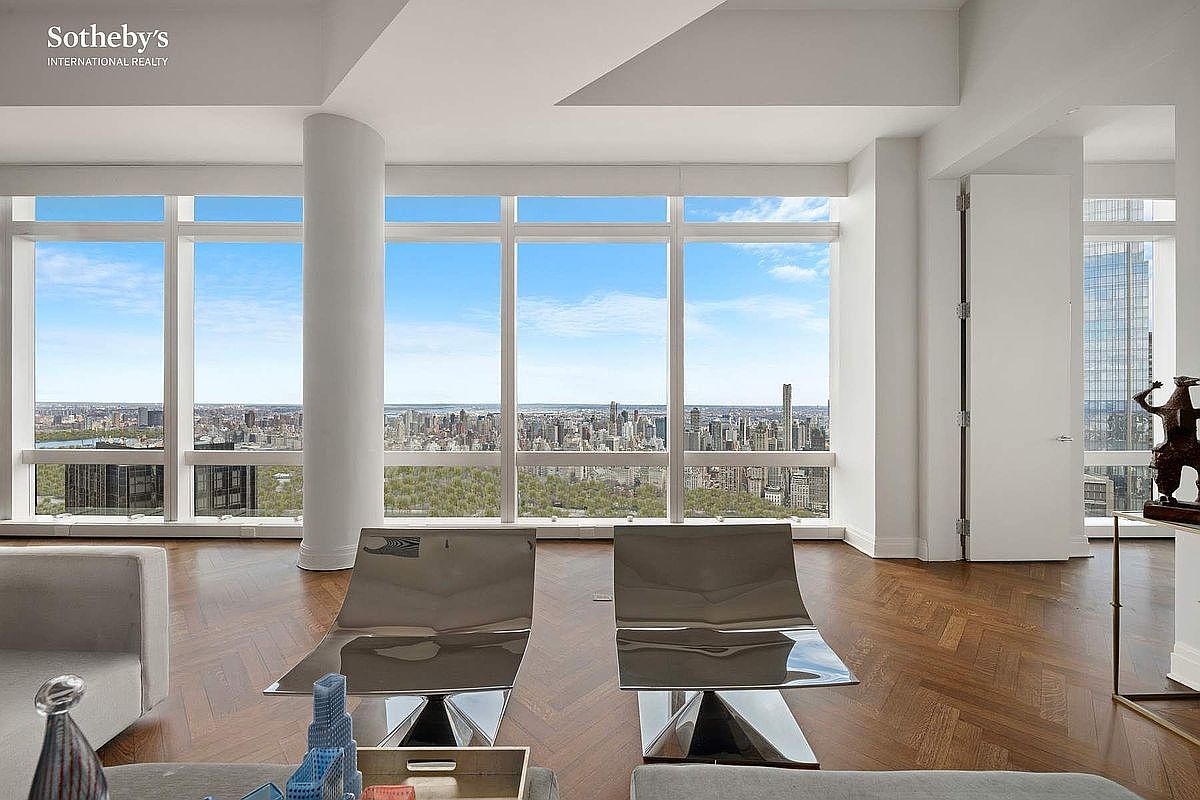 This sophisticated living room features floor-to-ceiling windows that offer a breathtaking, panoramic view of the city skyline and Central Park. The space is anchored by rich, dark herringbone hardwood floors and furnished with sleek, modern metallic chairs that reflect the natural light. The overall aesthetic is one of high-end luxury, emphasizing the expansive vistas and clean, minimalist architectural lines.