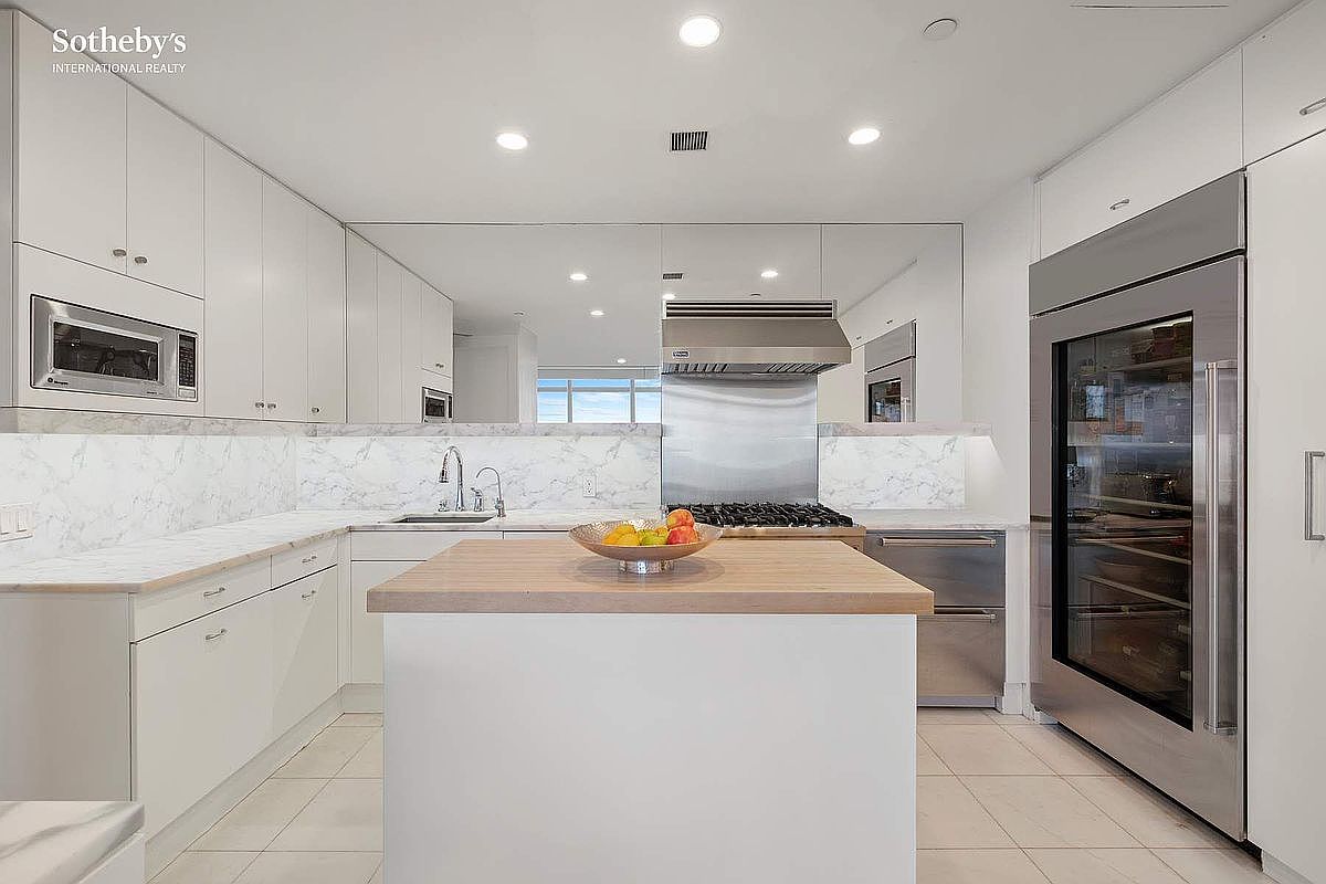 This sleek, modern kitchen features crisp white cabinetry, marble-patterned countertops, and high-end stainless steel appliances, including a large built-in refrigerator. A central island with a light wood countertop serves as a functional focal point, while a mirrored backsplash adds depth and reflects the bright, recessed lighting. The overall aesthetic is clean, minimalist, and sophisticated, offering a bright and airy atmosphere perfect for contemporary living.