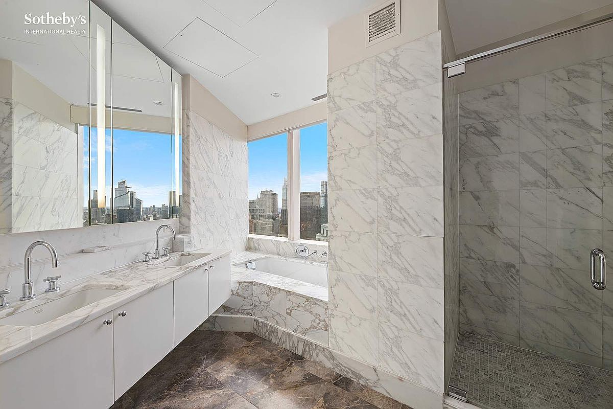 This luxurious primary bathroom features high-end marble tiling throughout the walls and a matching marble-clad soaking tub positioned next to floor-to-ceiling windows with panoramic city views. The space includes a sleek double vanity with modern fixtures and a spacious glass-enclosed walk-in shower. The overall aesthetic is sophisticated and contemporary, emphasizing natural light and premium materials.