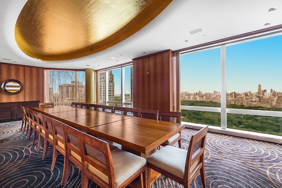 This sophisticated boardroom or executive office features a long, polished wooden conference table surrounded by matching chairs with woven cane accents. The room is defined by a striking, gold-leafed oval ceiling feature and floor-to-ceiling windows that provide a panoramic, elevated view of Central Park and the New York City skyline. The space is grounded by a dark, patterned carpet that adds a sense of luxury and professional gravitas to the interior.